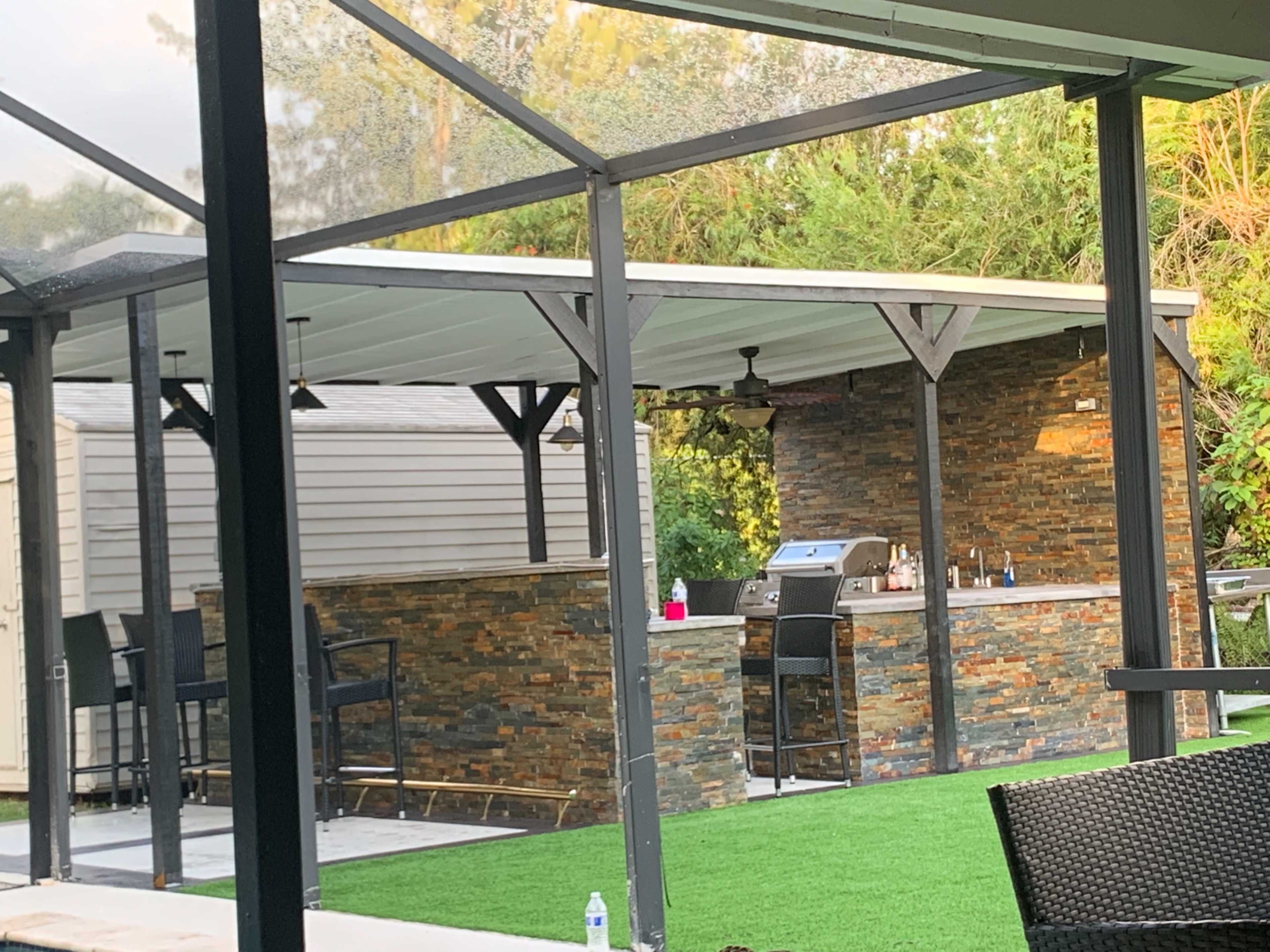 The image shows an outdoor kitchen area with a stone bar, a grill, and several tall bar stools under a white roof structure.