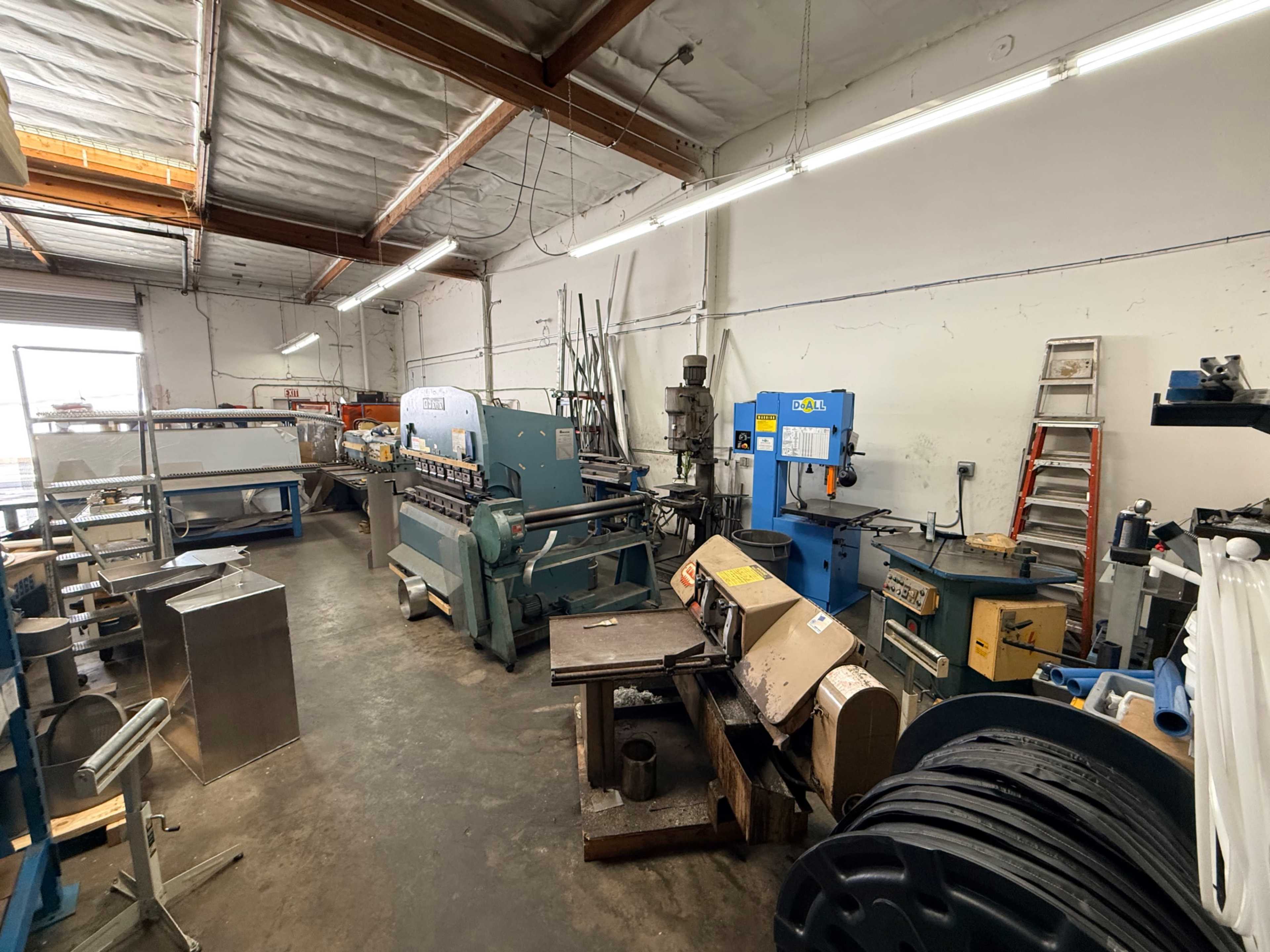 The image shows a workshop filled with various metalworking machines and tools, including a metal bending machine and a band saw, amid a cluttered space.
