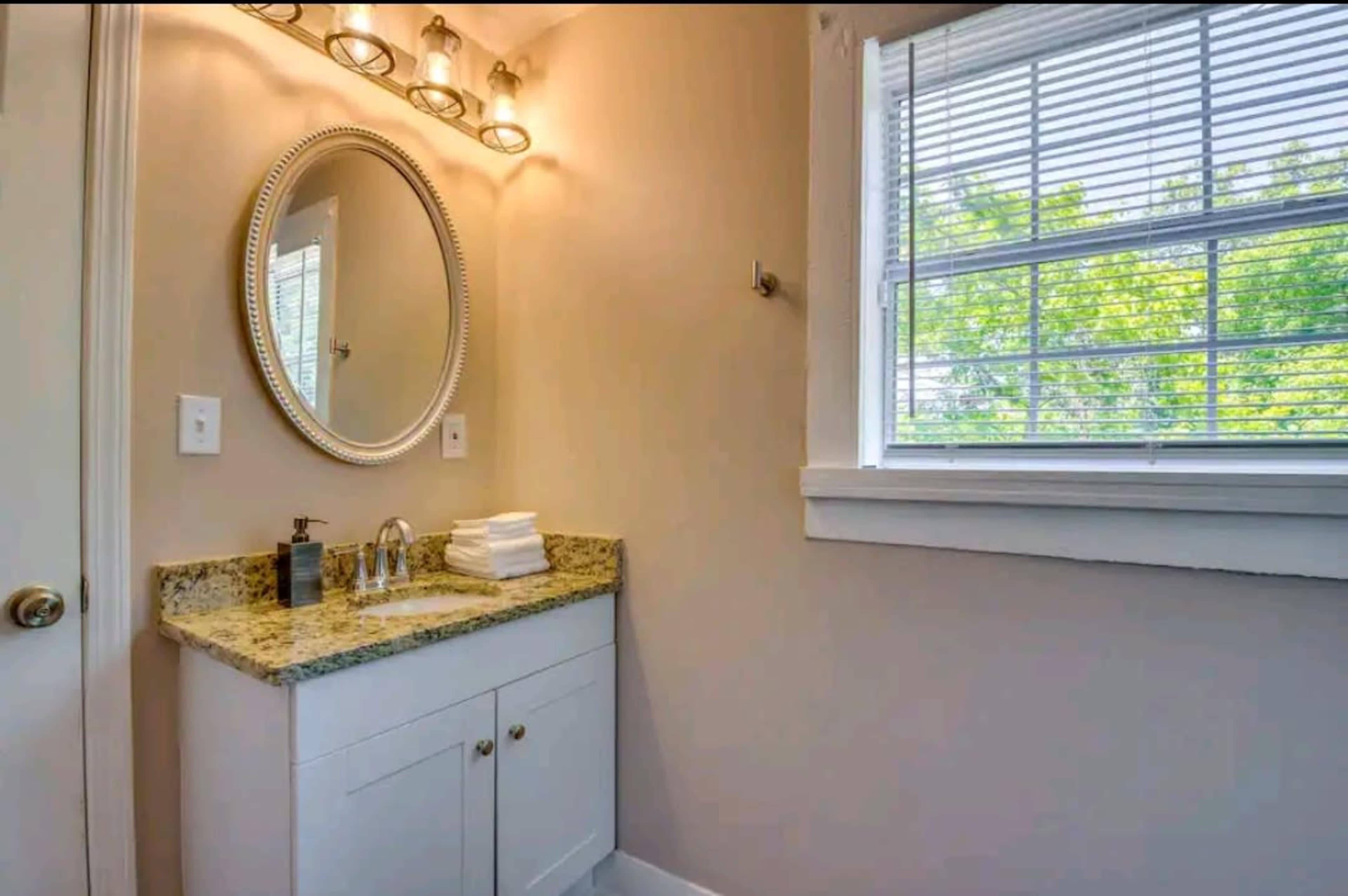 The image shows a bathroom with a mirror above a granite-topped vanity, featuring a faucet and neatly arranged toiletries, alongside a window with blinds.