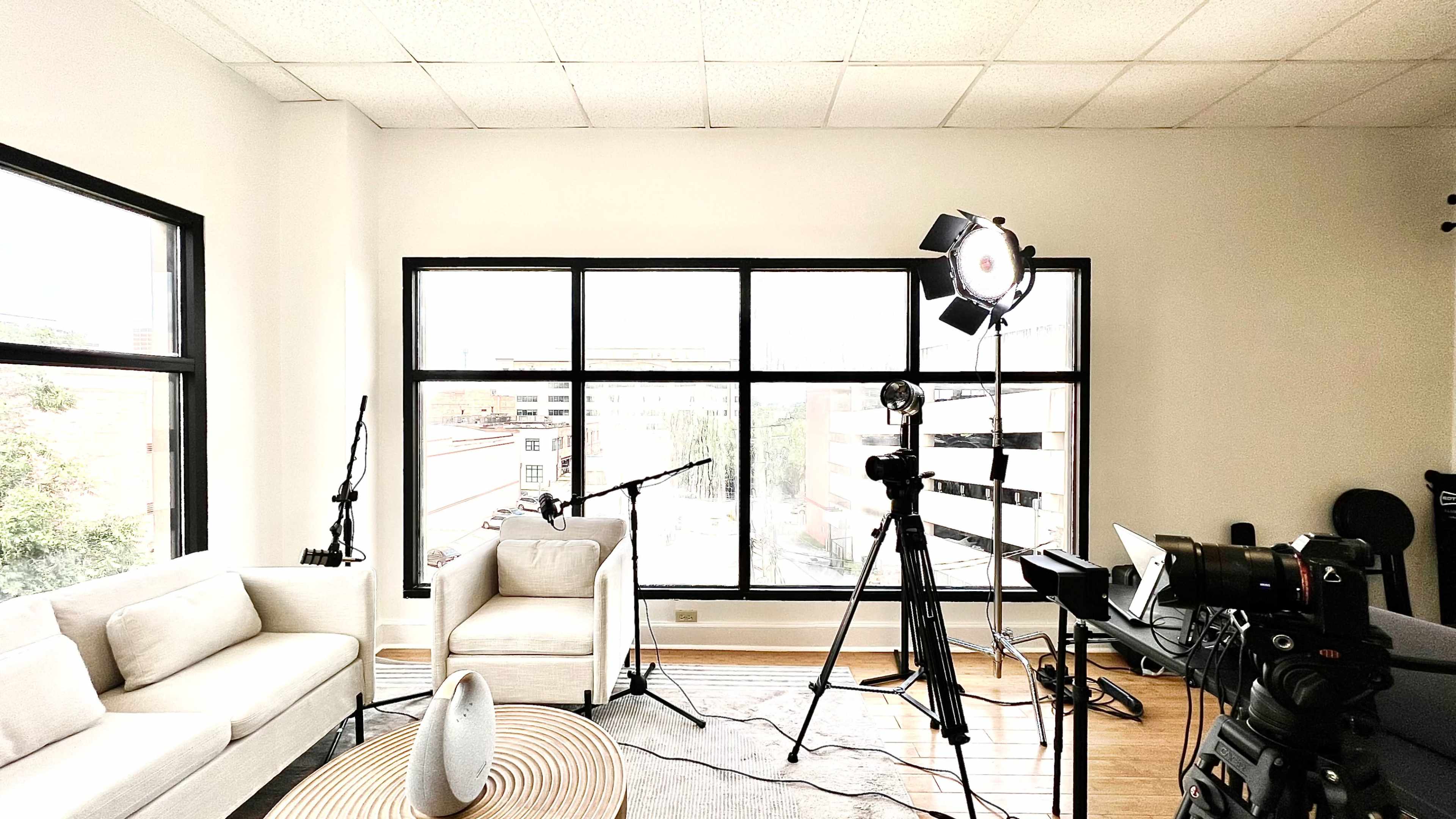 A film or photo studio setup featuring two chairs, a large window, lighting equipment, and a camera on a tripod.