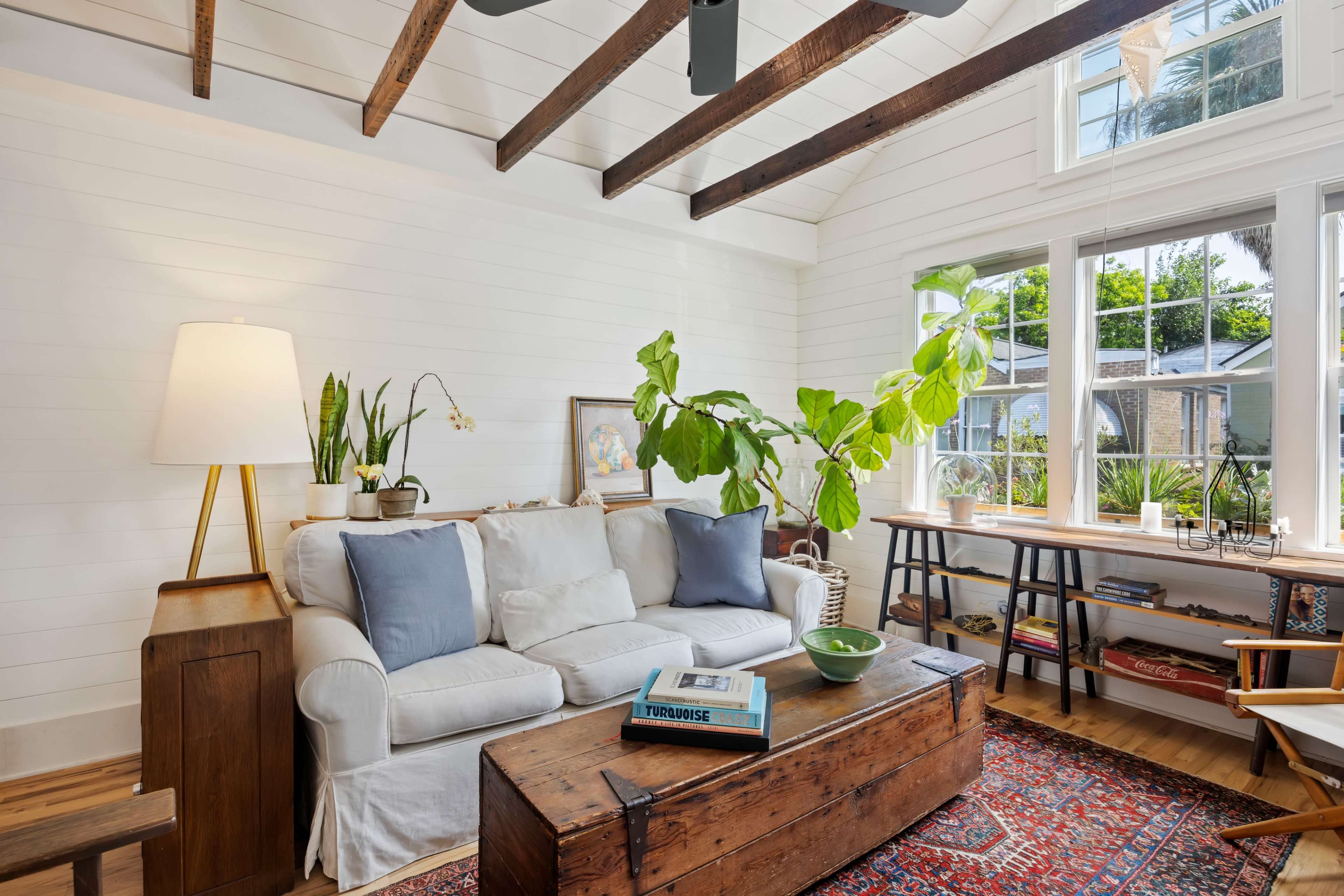 A bright living room features a white sofa with blue cushions, a wooden coffee table, and a large plant by the window.