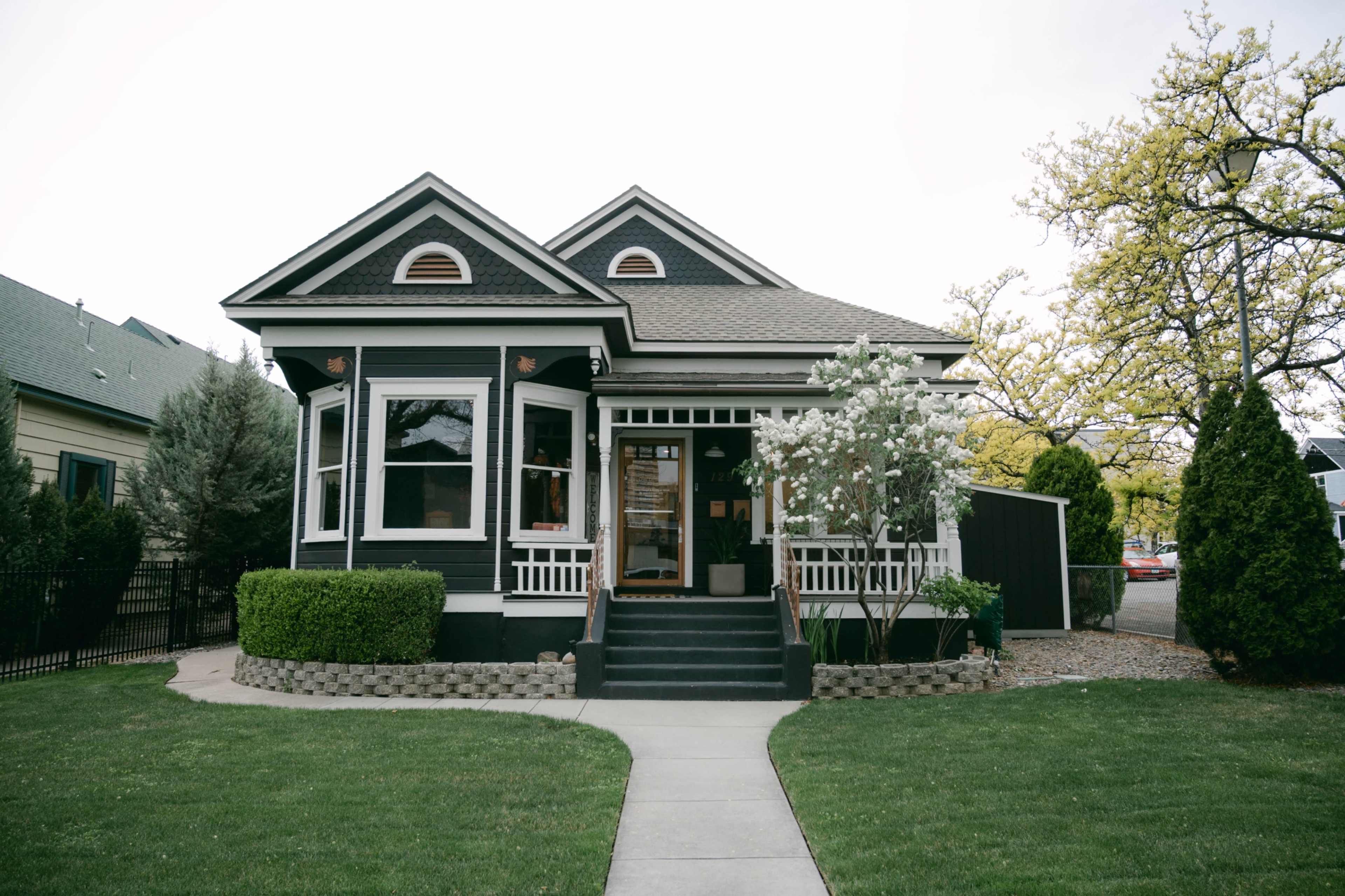 The image shows a black-painted house with a front porch, surrounded by well-maintained greenery and a pathway leading to the entrance.