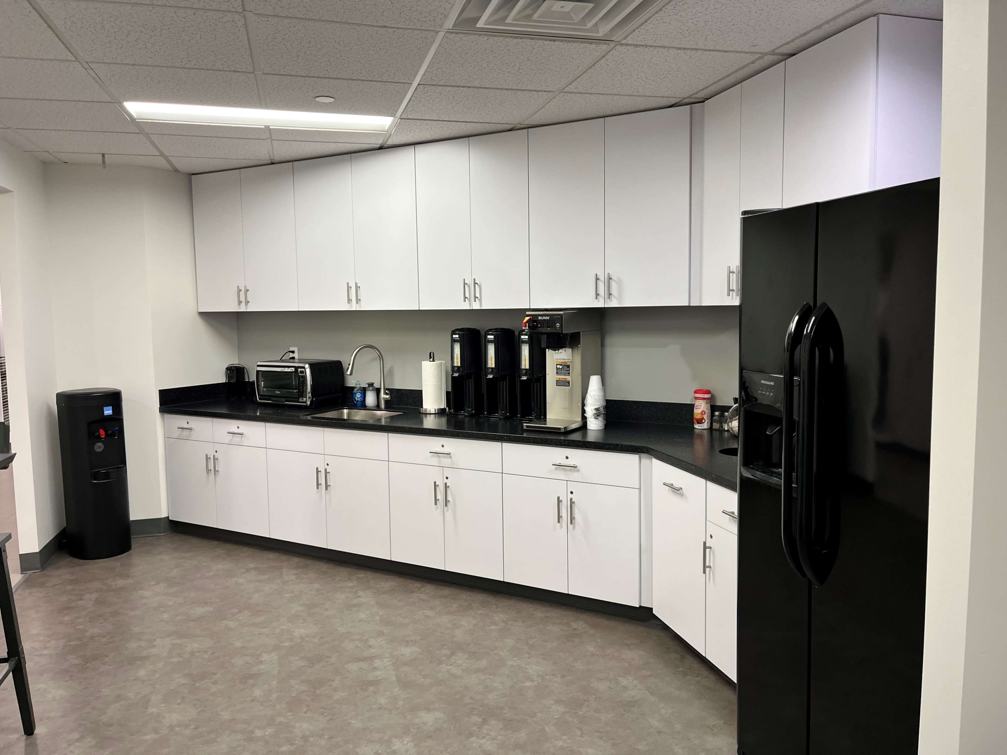The image shows a modern kitchen with white cabinets, a black refrigerator, an oven, and several coffee machines along a black countertop.
