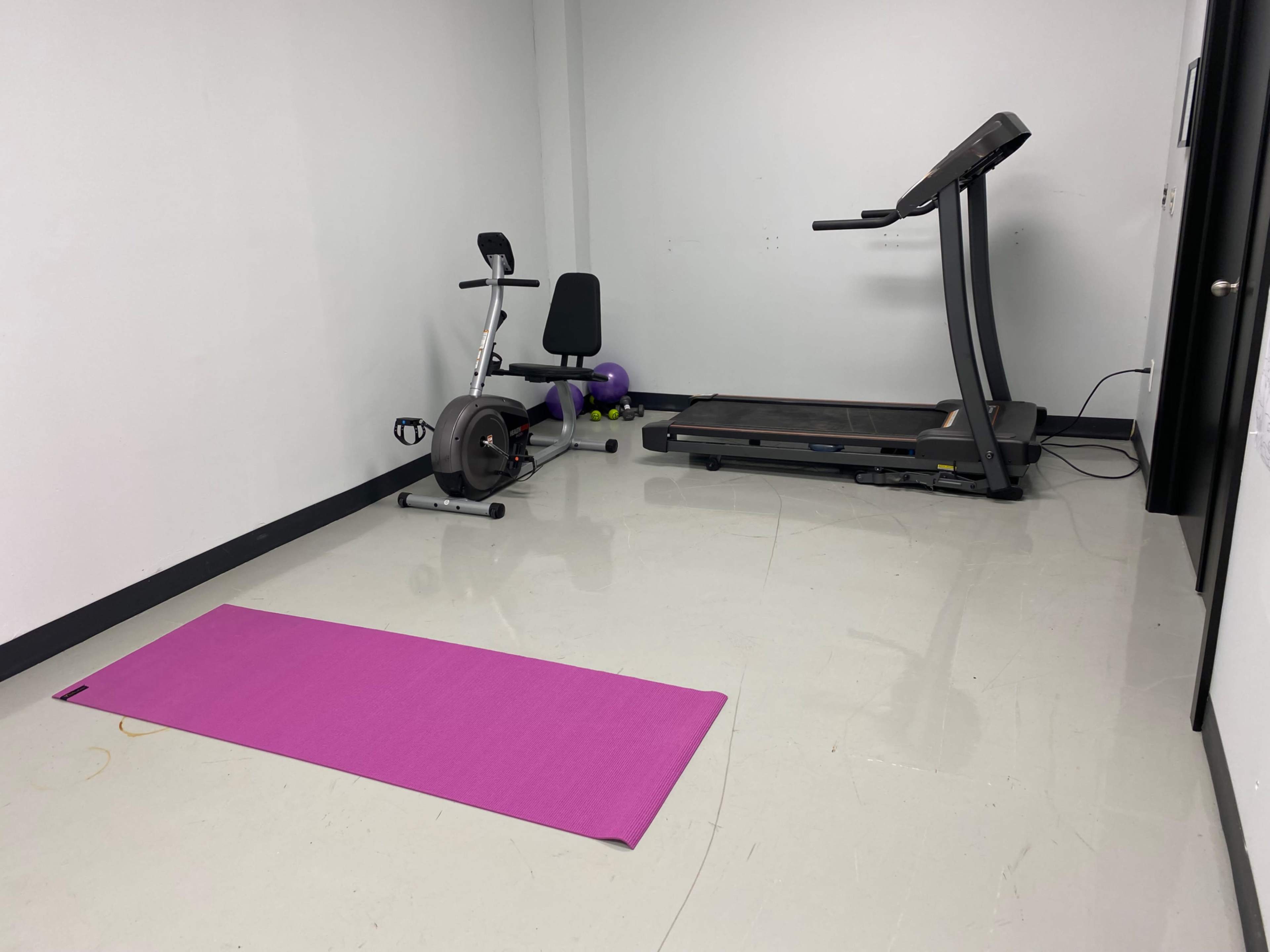 A small, minimalistic gym space featuring a purple exercise mat, a stationary bike, and a treadmill.