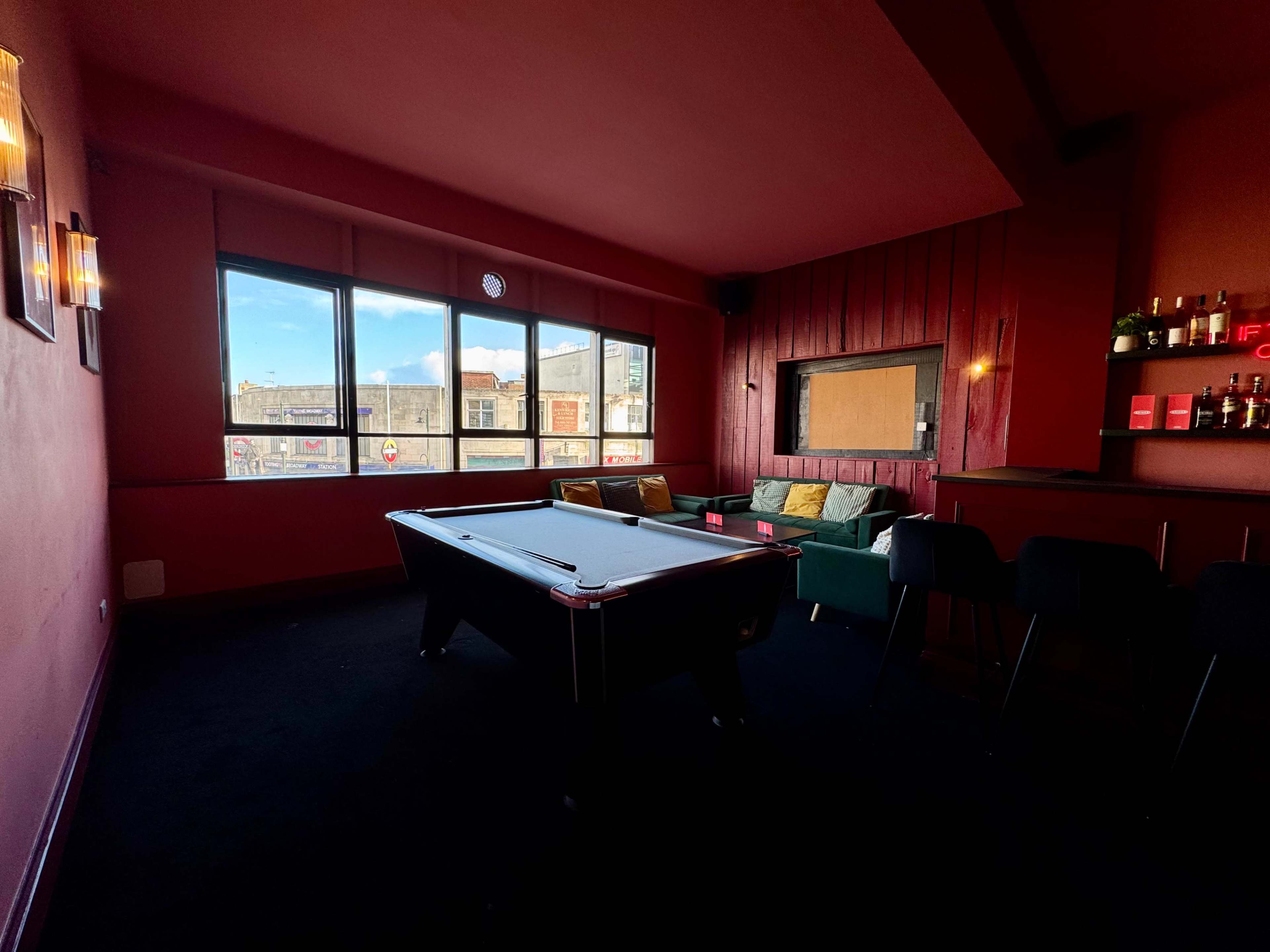 The image shows a pool table in a dimly lit room with red walls, large windows letting in natural light, and a bar area in the background.