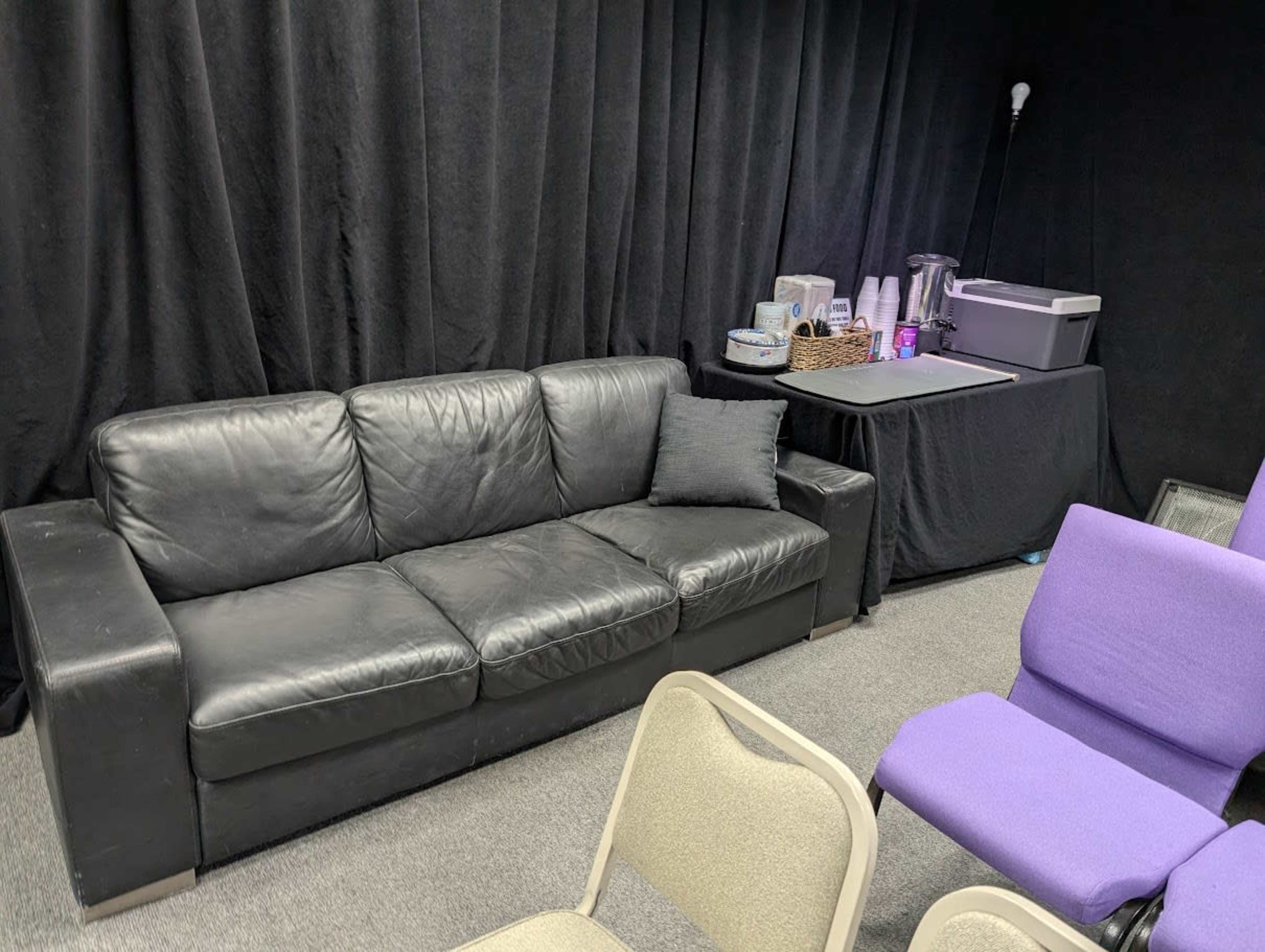 The image shows a black leather sofa positioned in a room with dark curtains, a table covered with a black cloth, and several chairs arranged nearby.