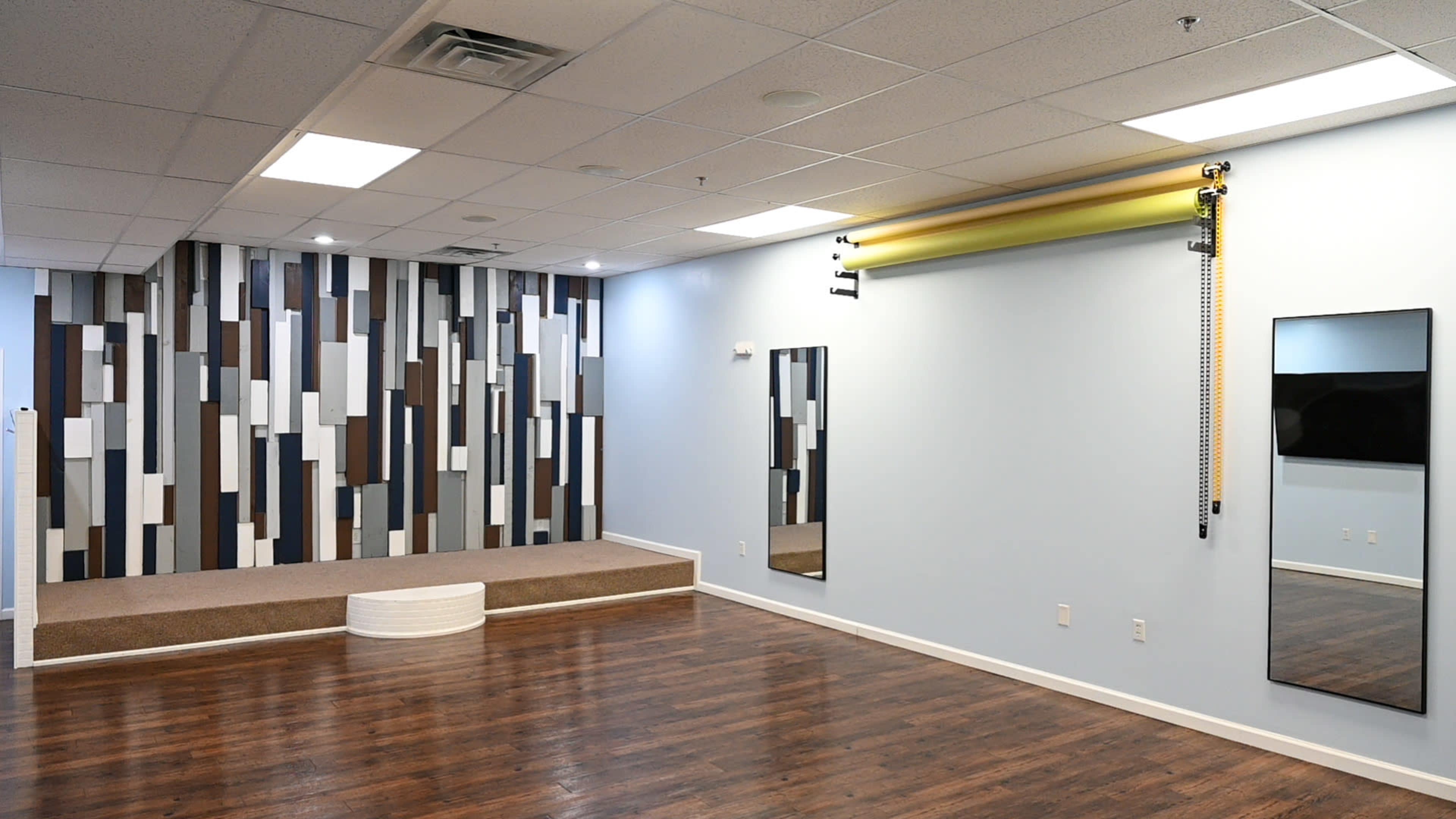 A spacious room with a hardwood floor, featuring a decorative wall of vertical stripes, two mirrors, and a small raised platform.