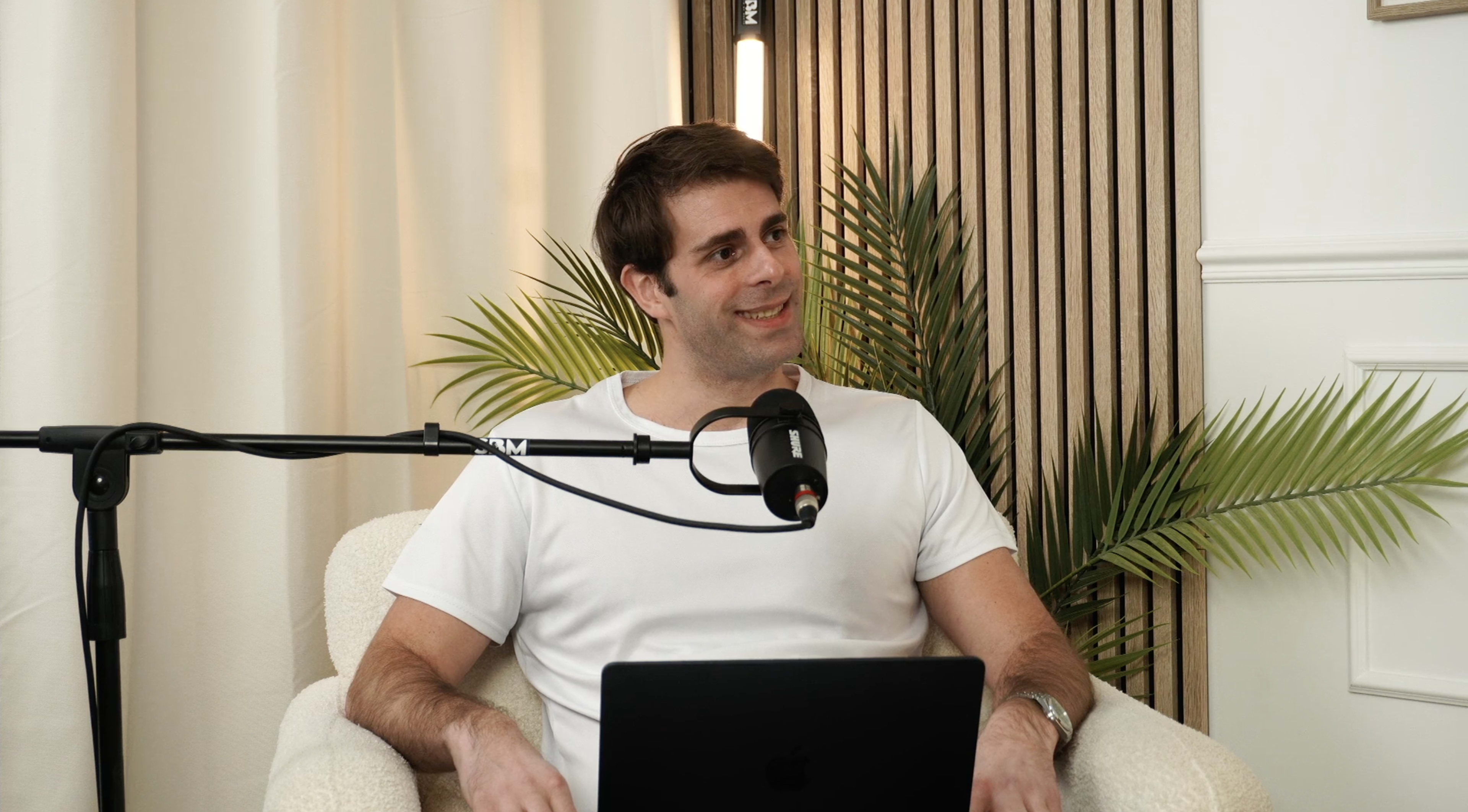 A man in a white shirt sits in a chair with a laptop and a microphone, surrounded by indoor plants and wooden paneling.