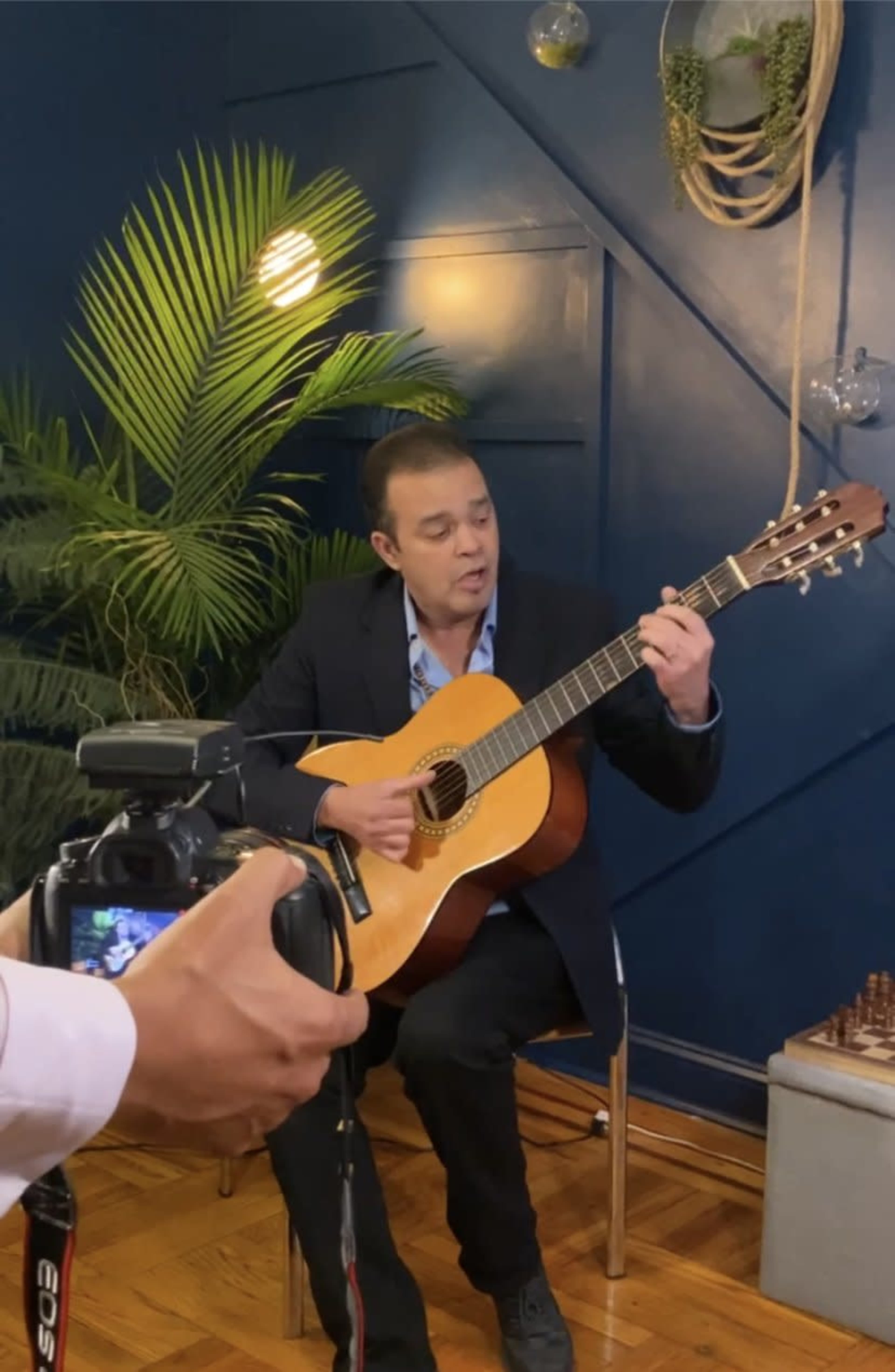 A man in a suit plays an acoustic guitar while sitting on a chair, with a camera set up in front of him.