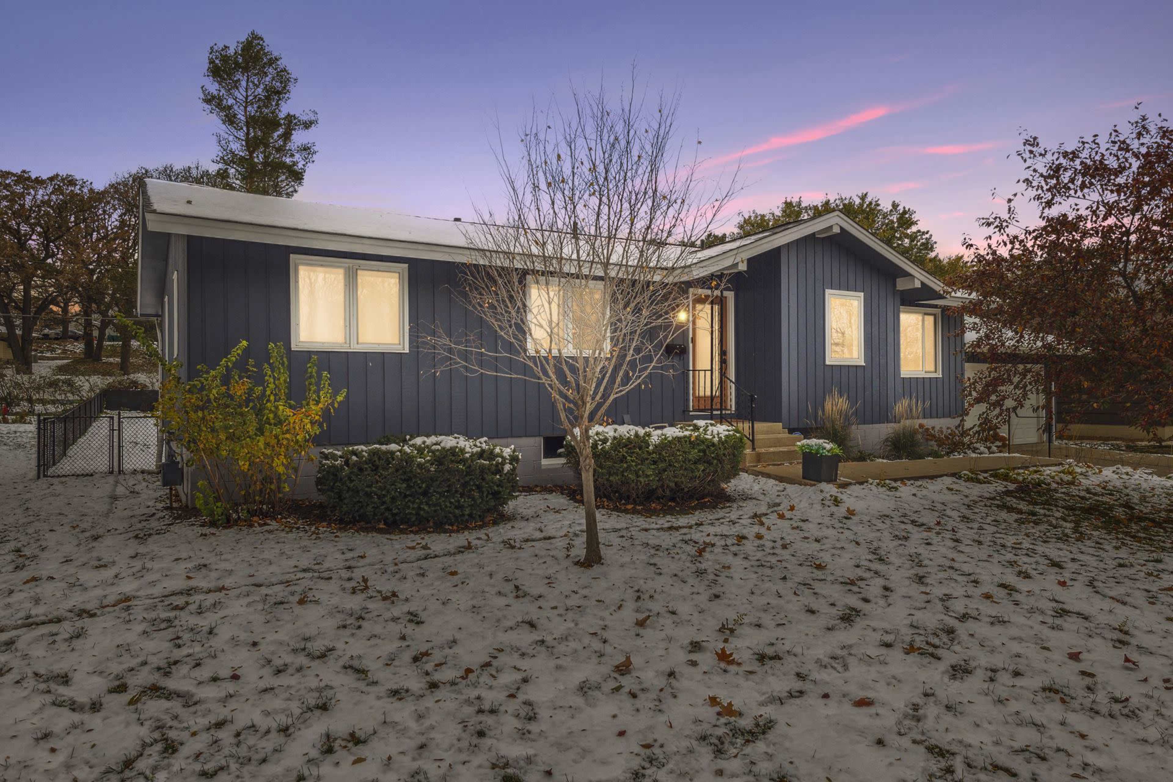 A blue single-story house with white accents is surrounded by a snow-covered yard and bare trees at dusk.