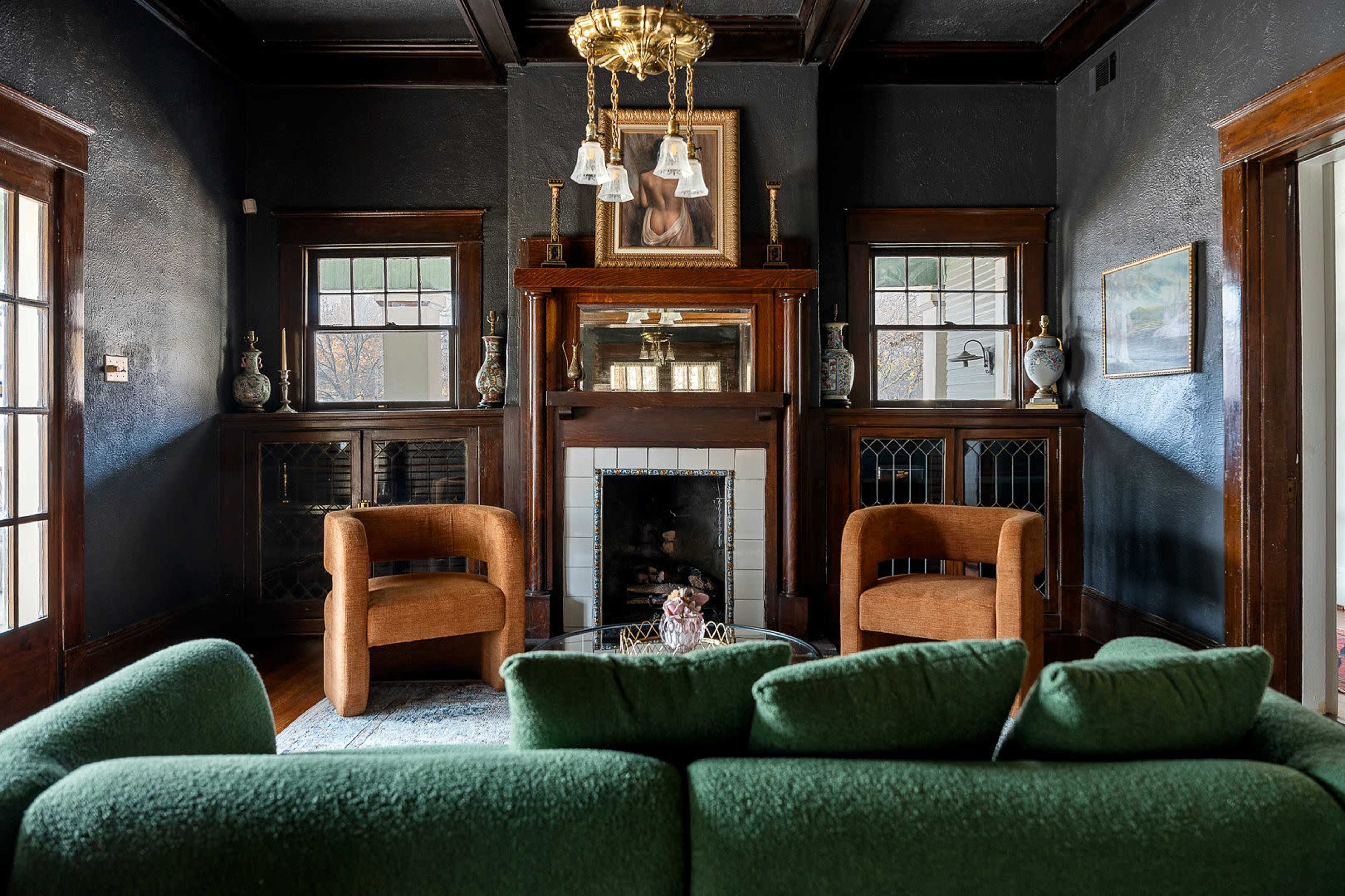 The image shows a richly decorated living room featuring two brown armchairs and a green sofa, with a fireplace and artwork on the walls.