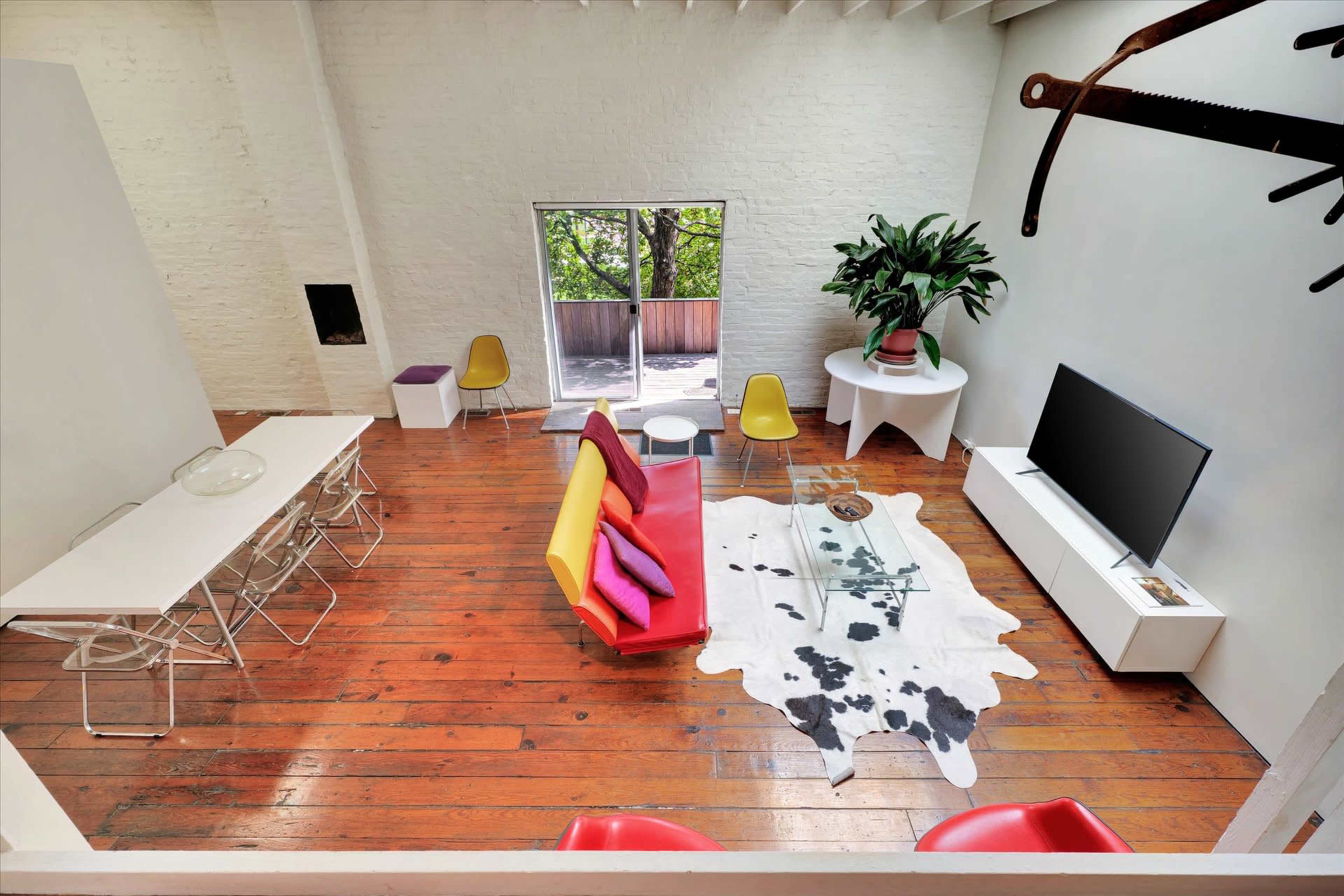 The image shows a bright, modern living space with wooden floors, a red and yellow sofa, a dining table, a television, and a large plant.
