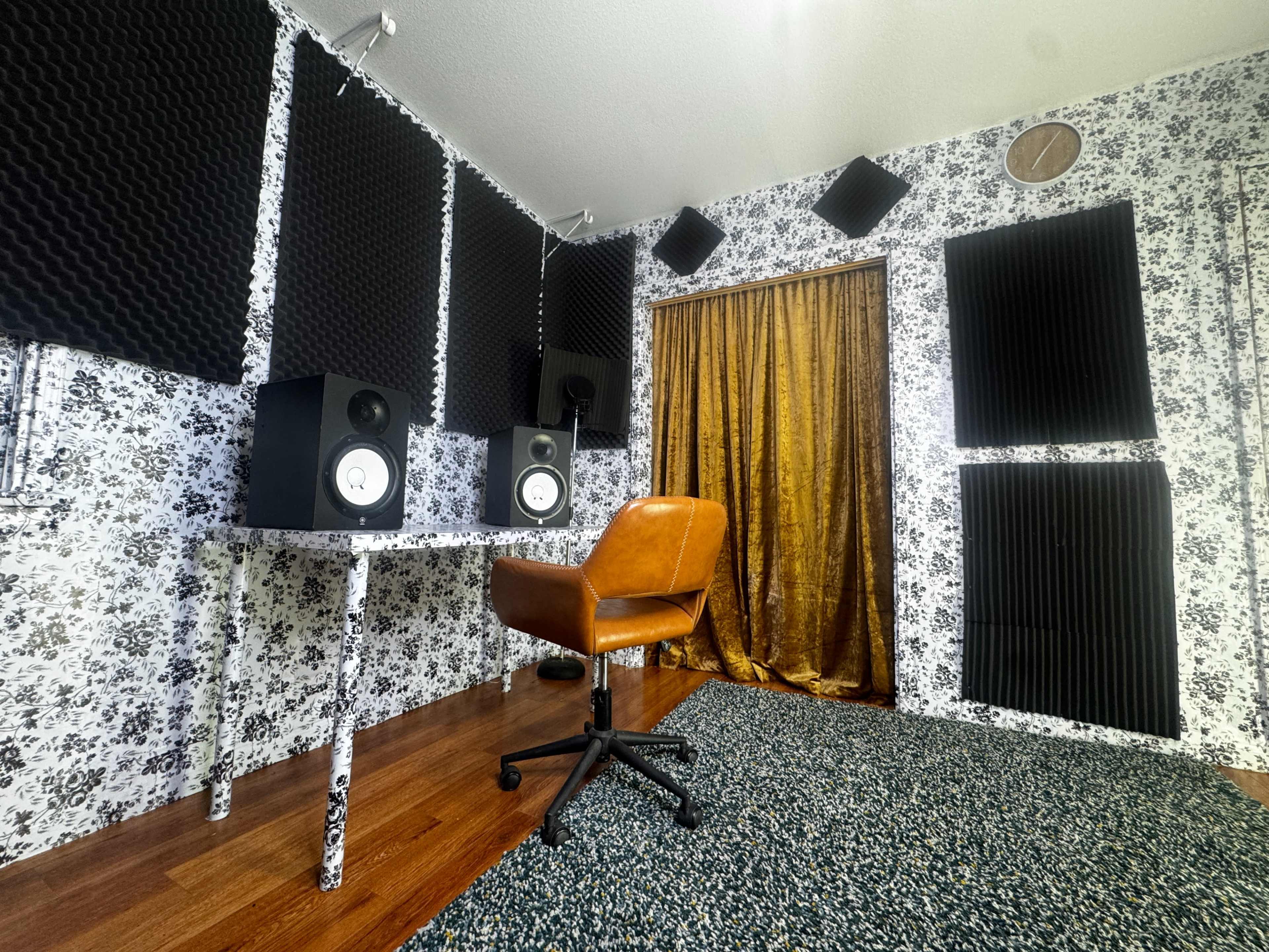 The image shows a small audio recording studio with soundproofed walls, a desk with studio monitors, and a vintage leather chair facing a curtain.