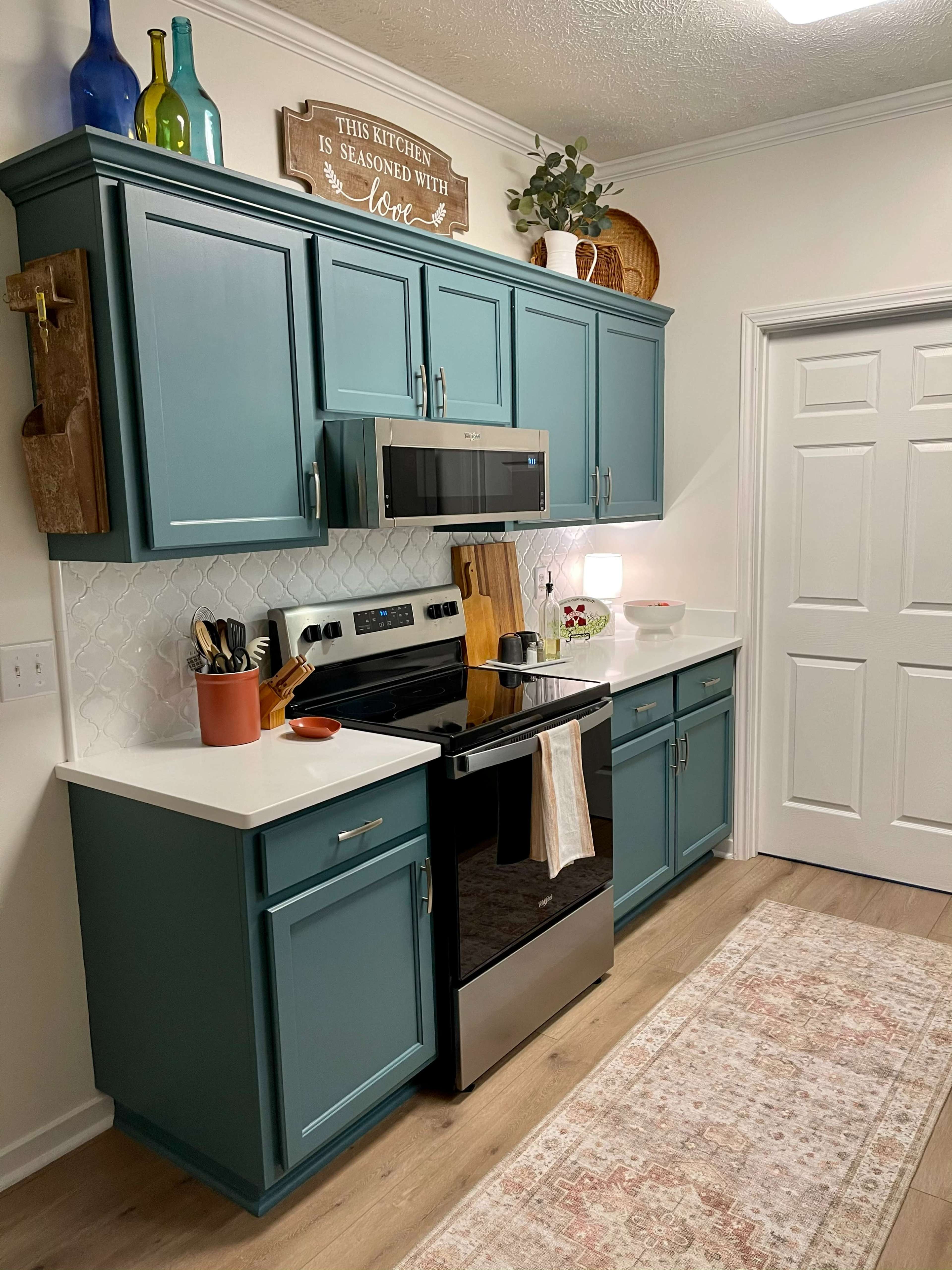 A tidy kitchen with teal cabinets, a stainless steel stove, and a light-colored rug on the floor.