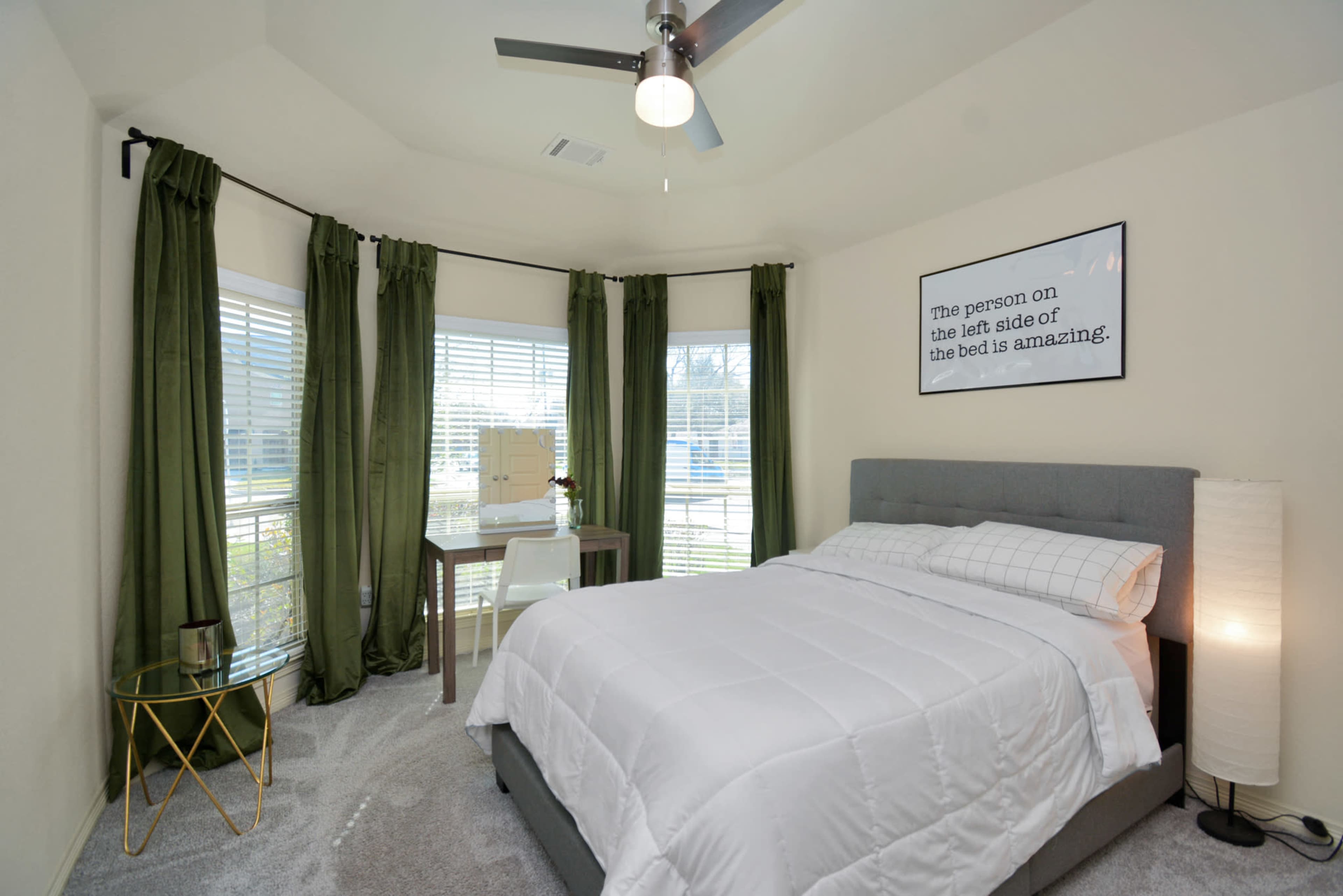 A neatly arranged bedroom features a gray bed, a small desk, large windows with green curtains, and a ceiling fan.