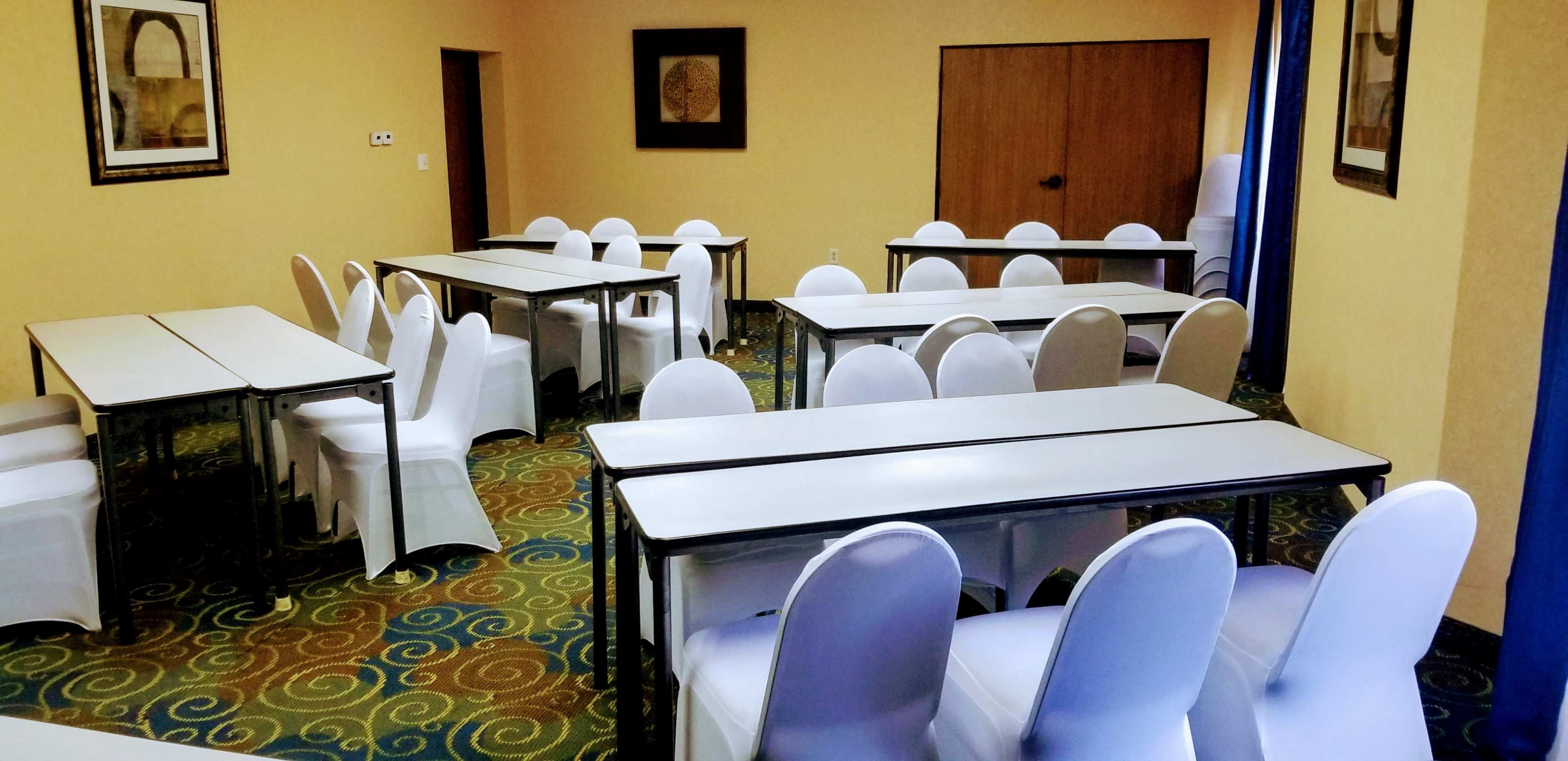 The image shows a conference room with several rectangular tables arranged in rows, surrounded by white chairs on a patterned carpet.