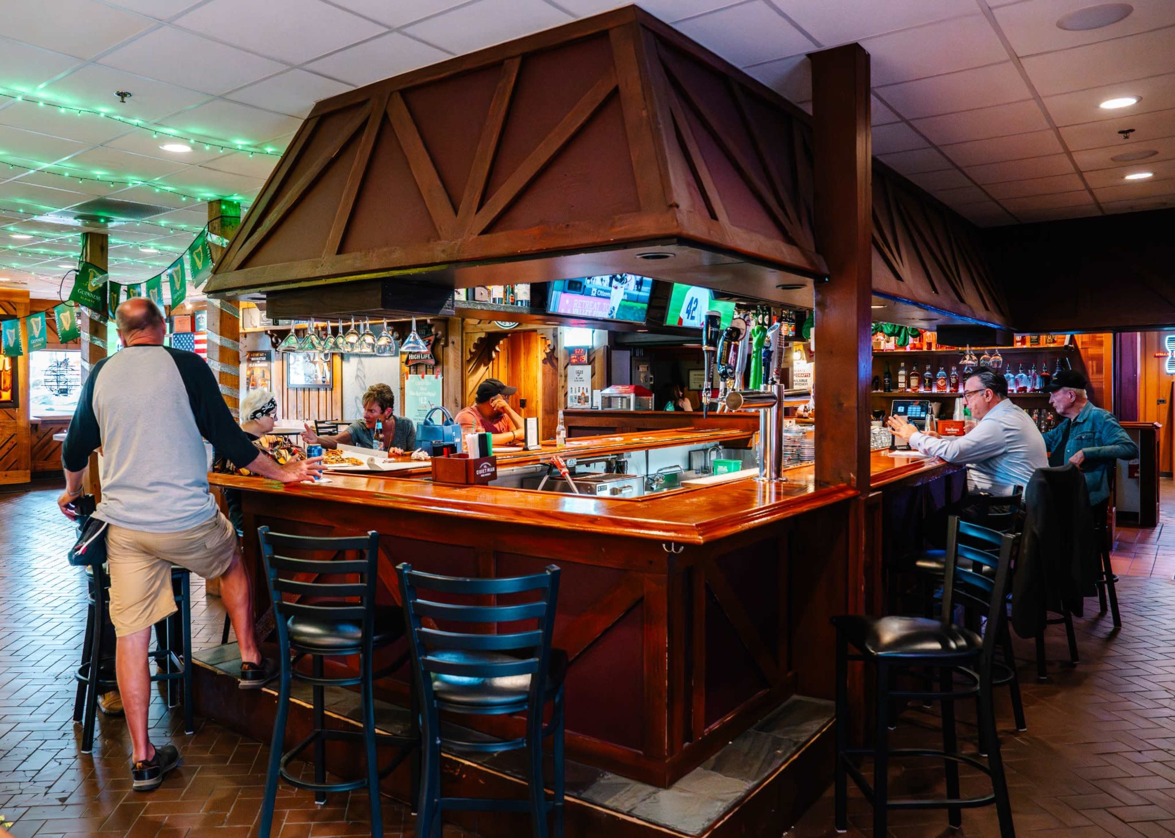 The image shows a bar area inside a pub with several patrons sitting and standing around, engaged in conversation and enjoying drinks.