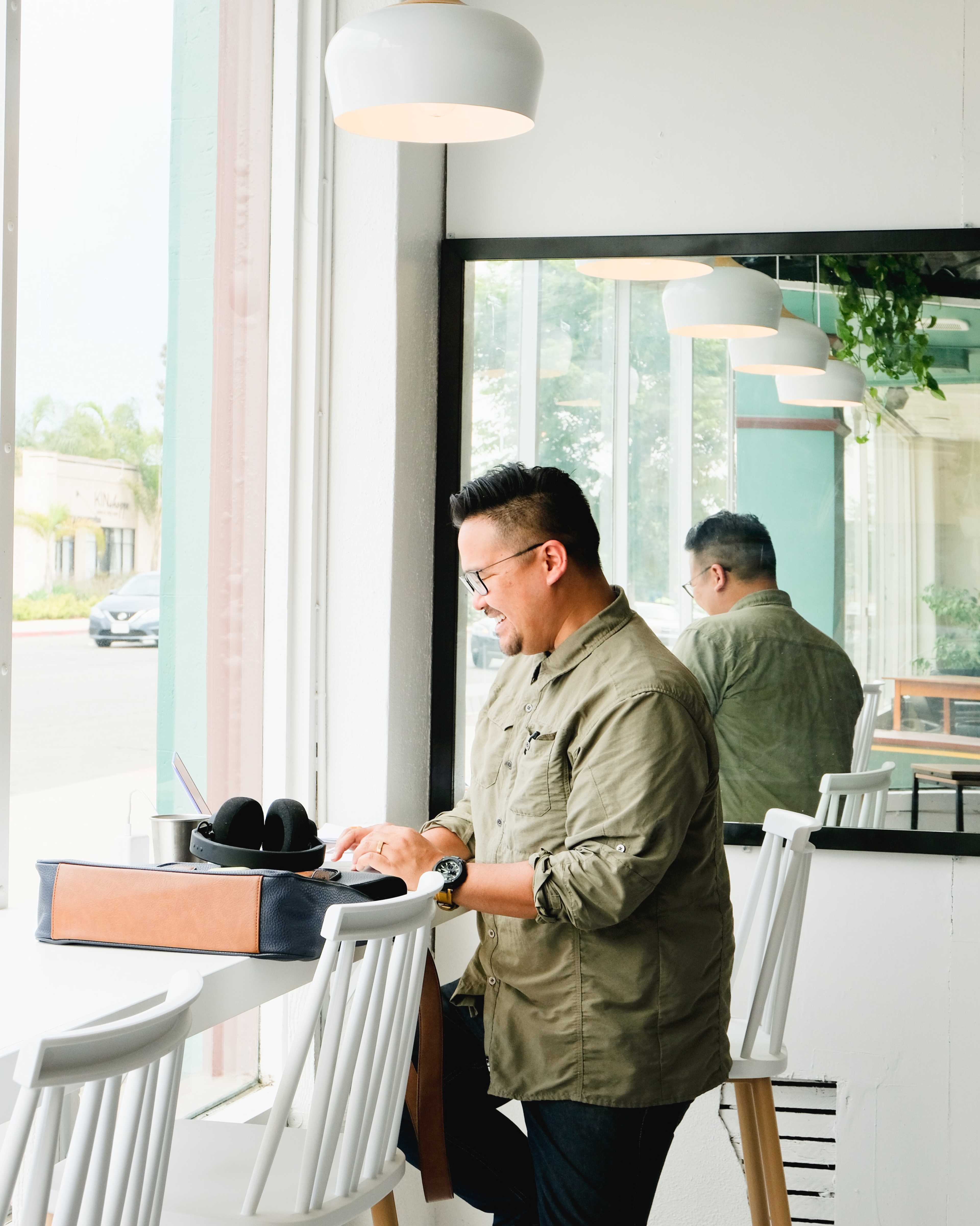 A man in an olive green shirt works on a laptop at a counter near a large window, with a mirror and a plant in the background.