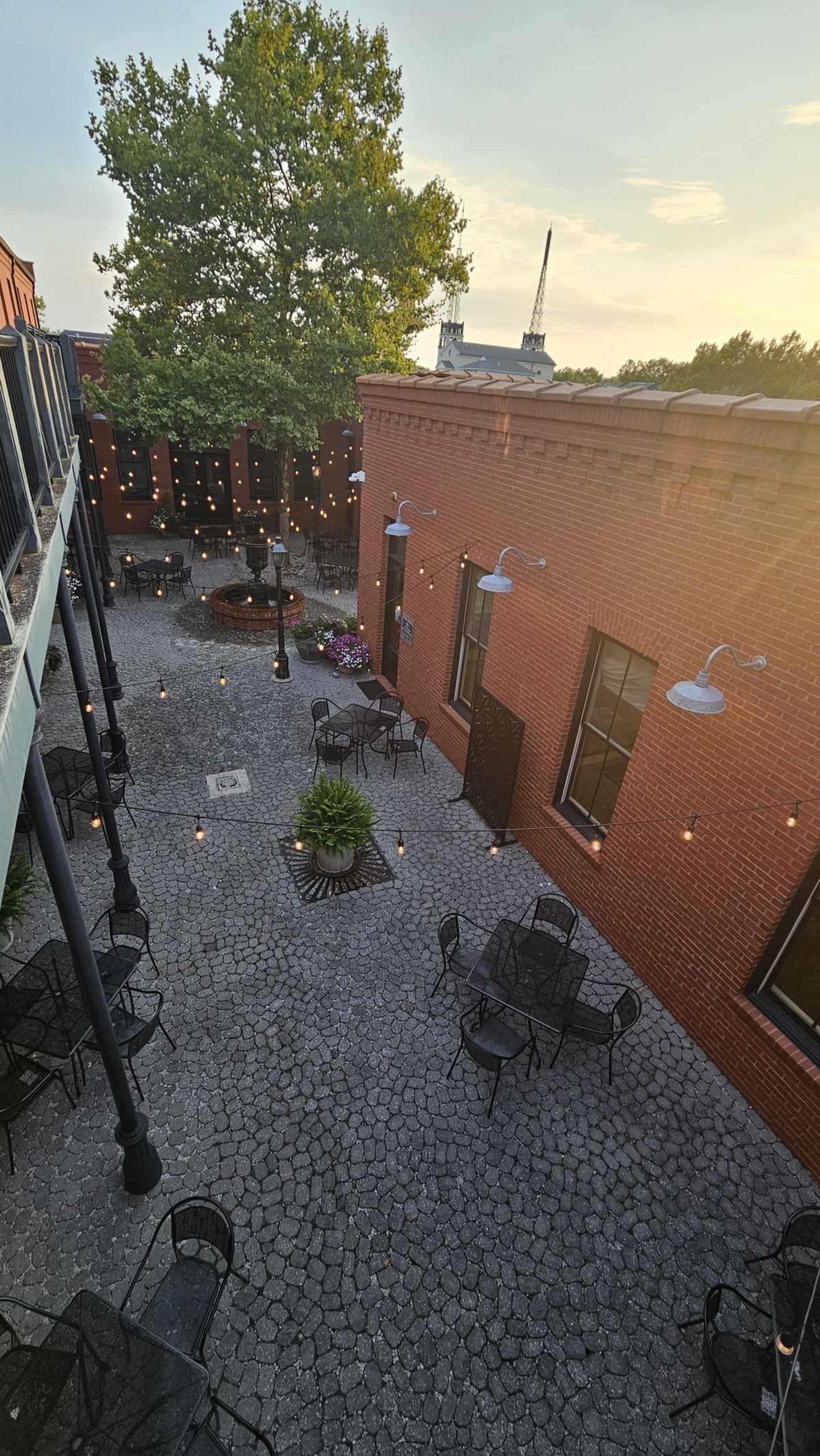 The image shows a courtyard with several round tables and chairs, surrounded by brick walls and illuminated by string lights.