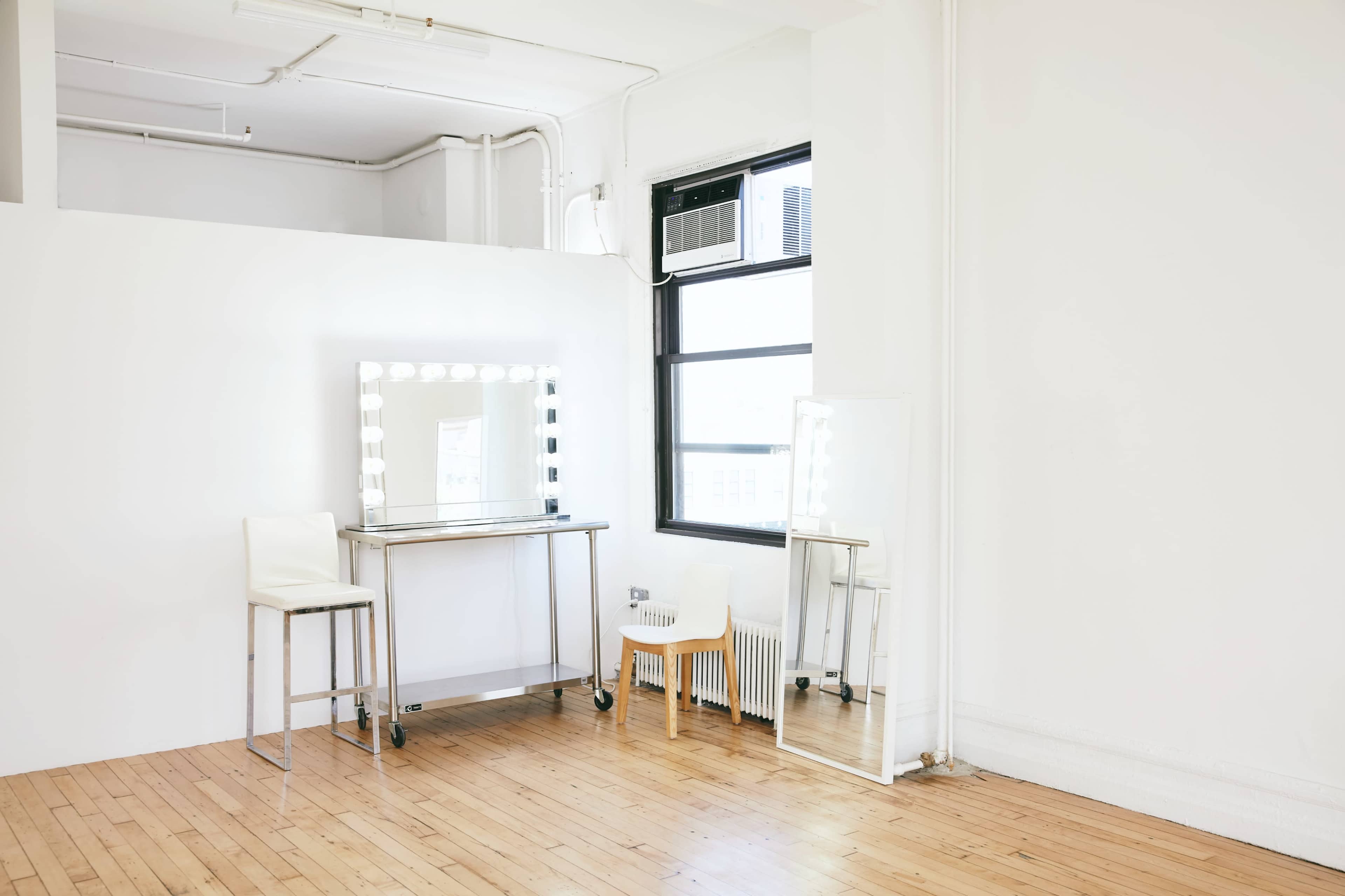 The image shows a minimalist room featuring a large illuminated mirror on a metal table, a tall mirror, and a wooden floor.