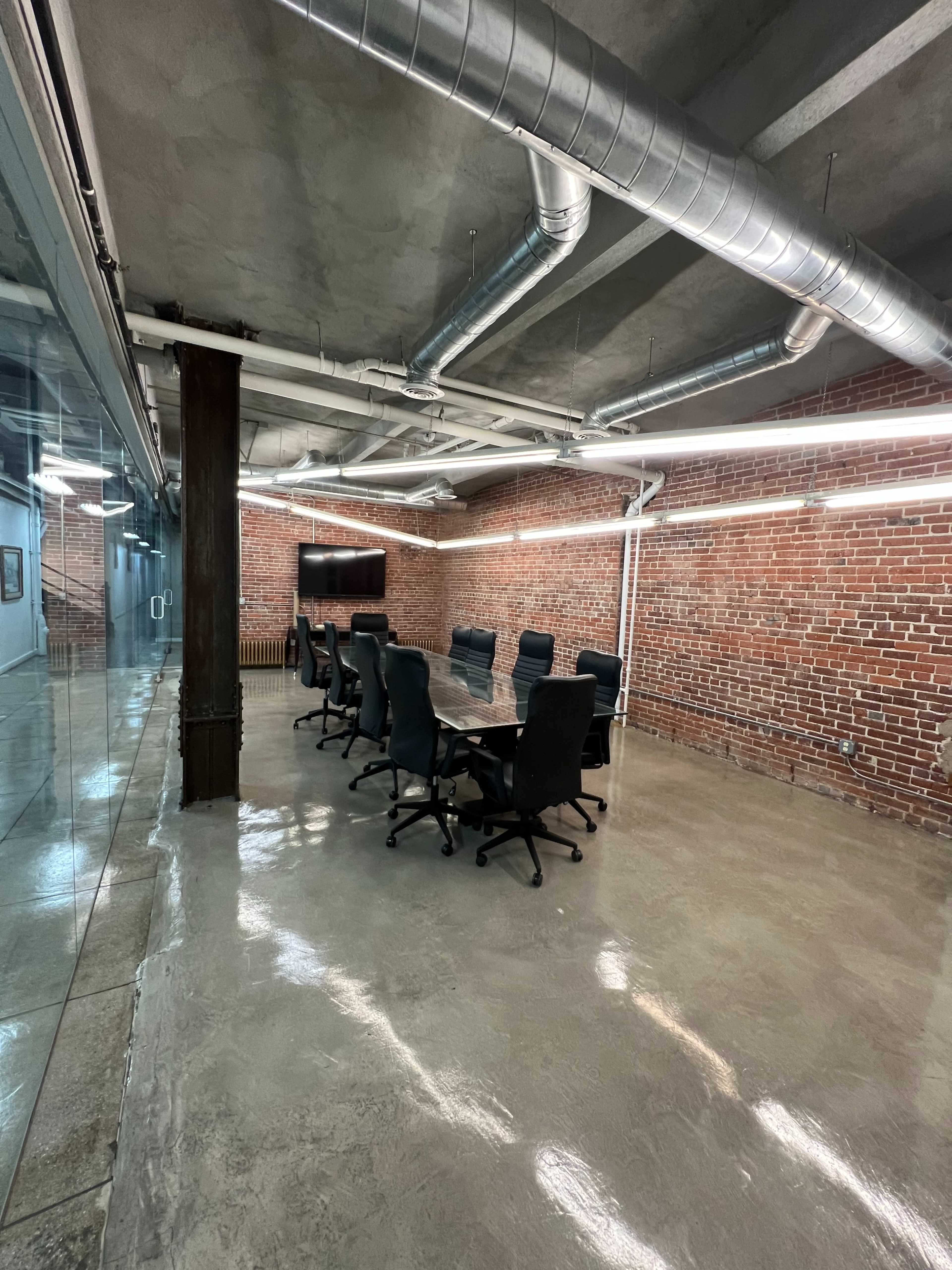 A conference room features a large table surrounded by black chairs, with exposed brick walls and industrial ductwork overhead.