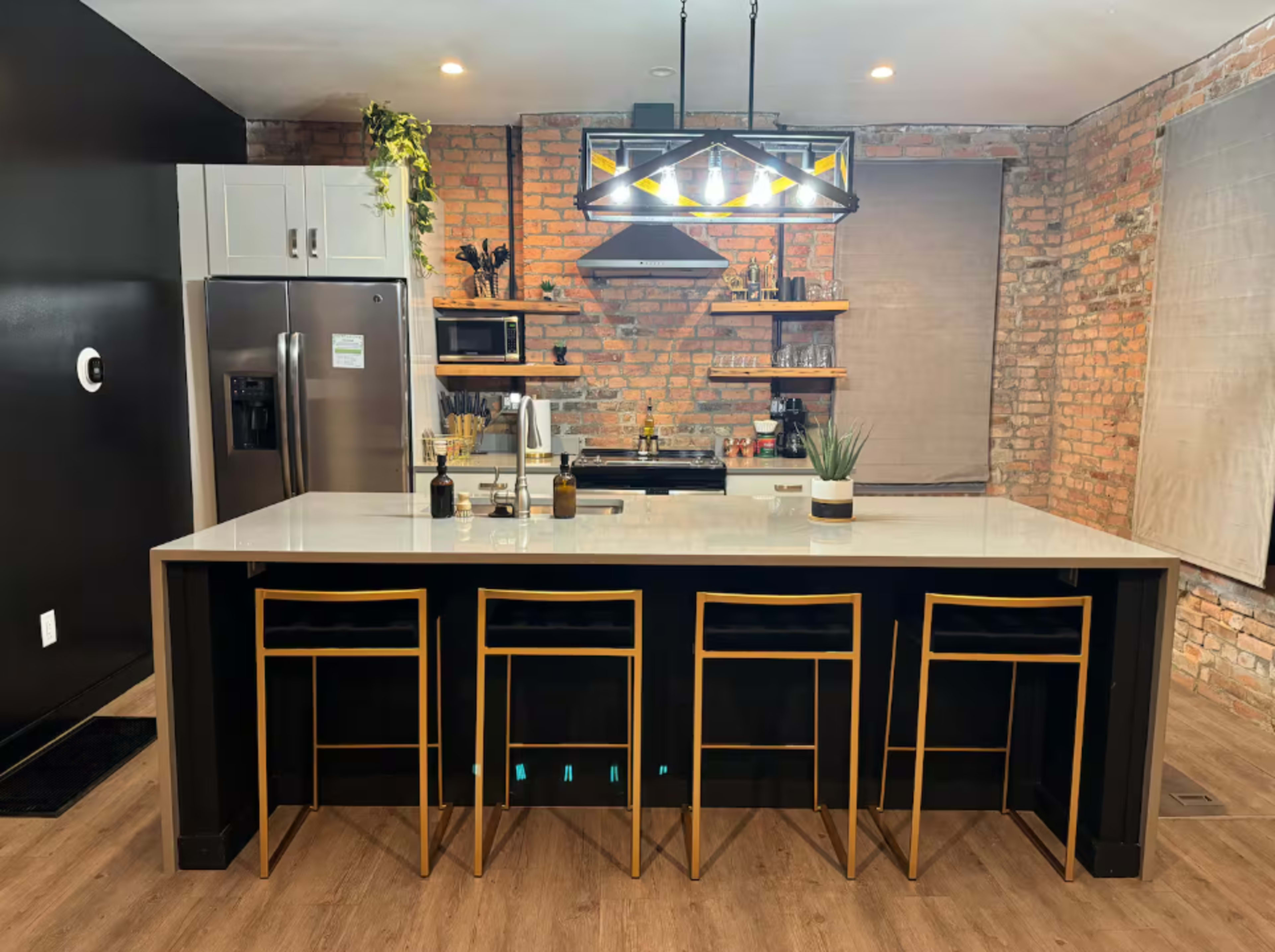 The kitchen features exposed brick walls, a large island with seating, stainless steel appliances, and modern lighting fixtures.