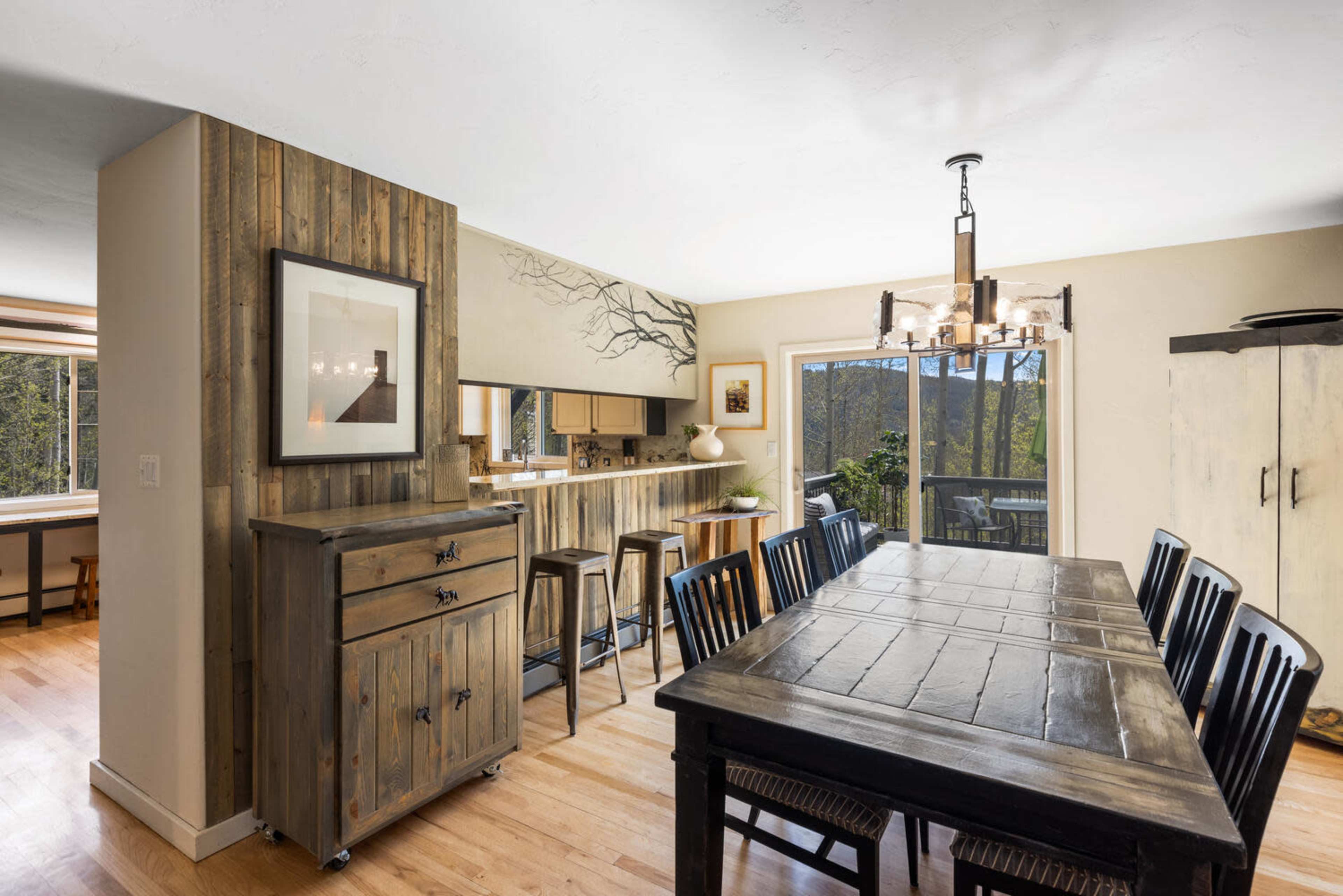The image shows a bright dining area with a wooden table and black chairs, adjacent to a kitchen with bar seating and large windows overlooking a natural landscape.