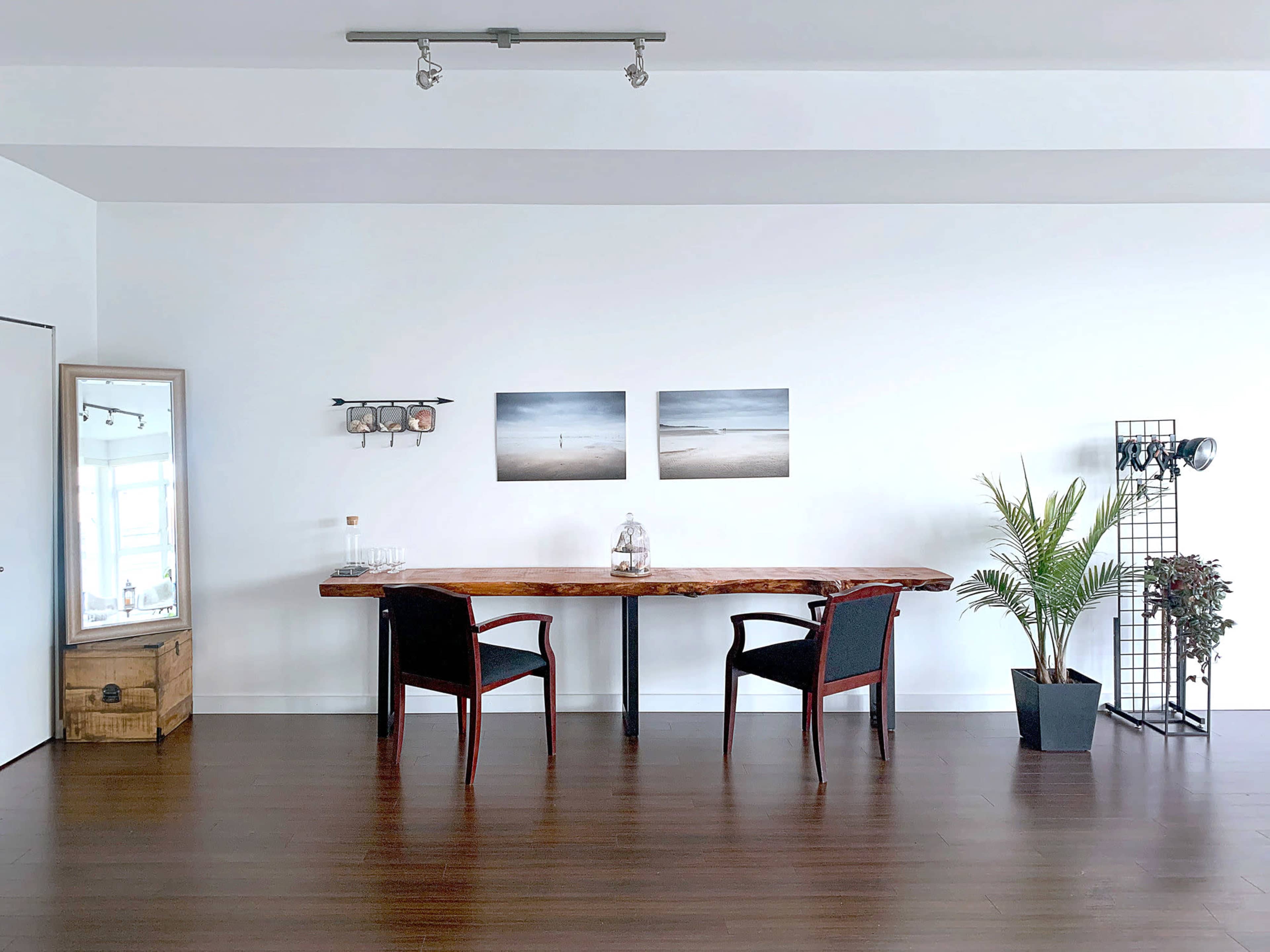 A minimalist room with a wooden dining table, two chairs, a large mirror, decorative plants, and two landscape photographs on the wall.