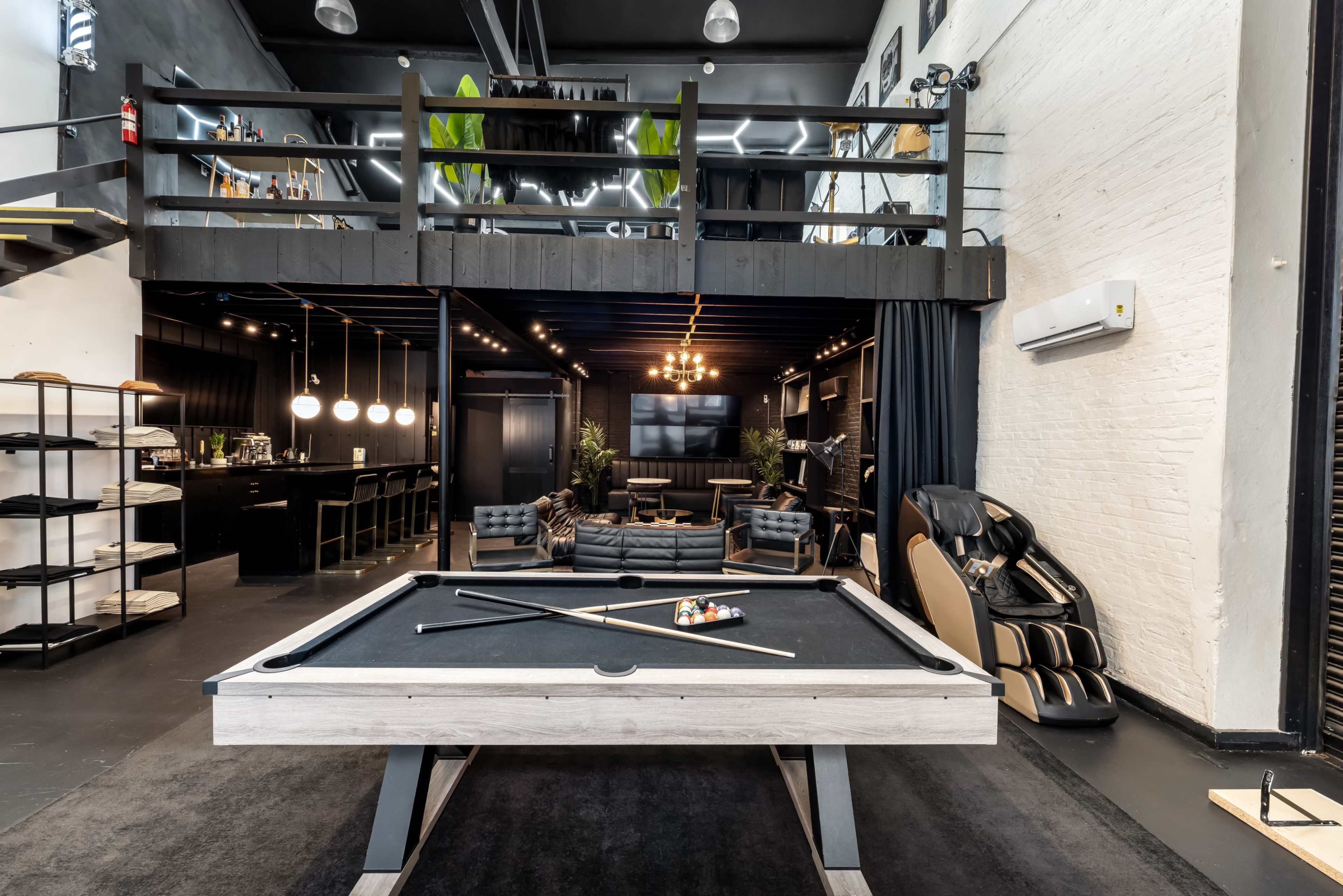 The image shows a modern lounge area featuring a black pool table in the foreground, with a spacious open layout that includes seating areas and a stylish bar on the upper level.