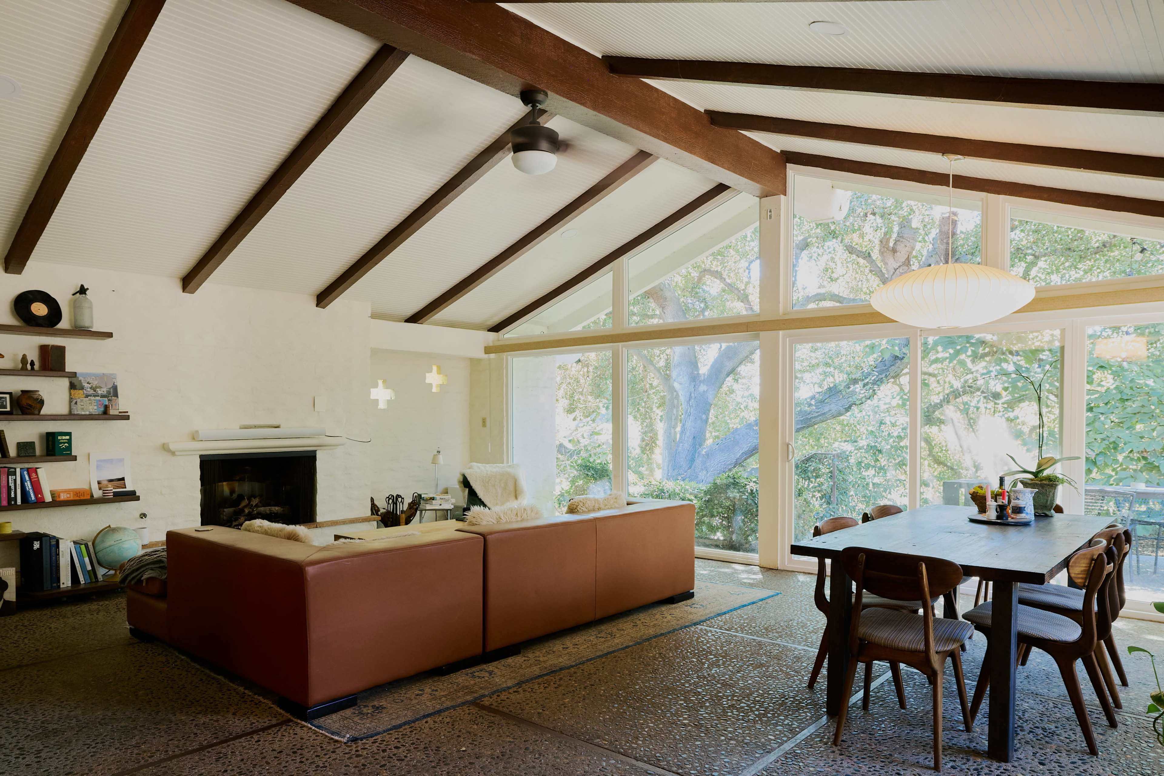 A spacious living area with a brown sectional sofa, a wooden dining table, and large windows overlooking a tree-filled outdoor space.