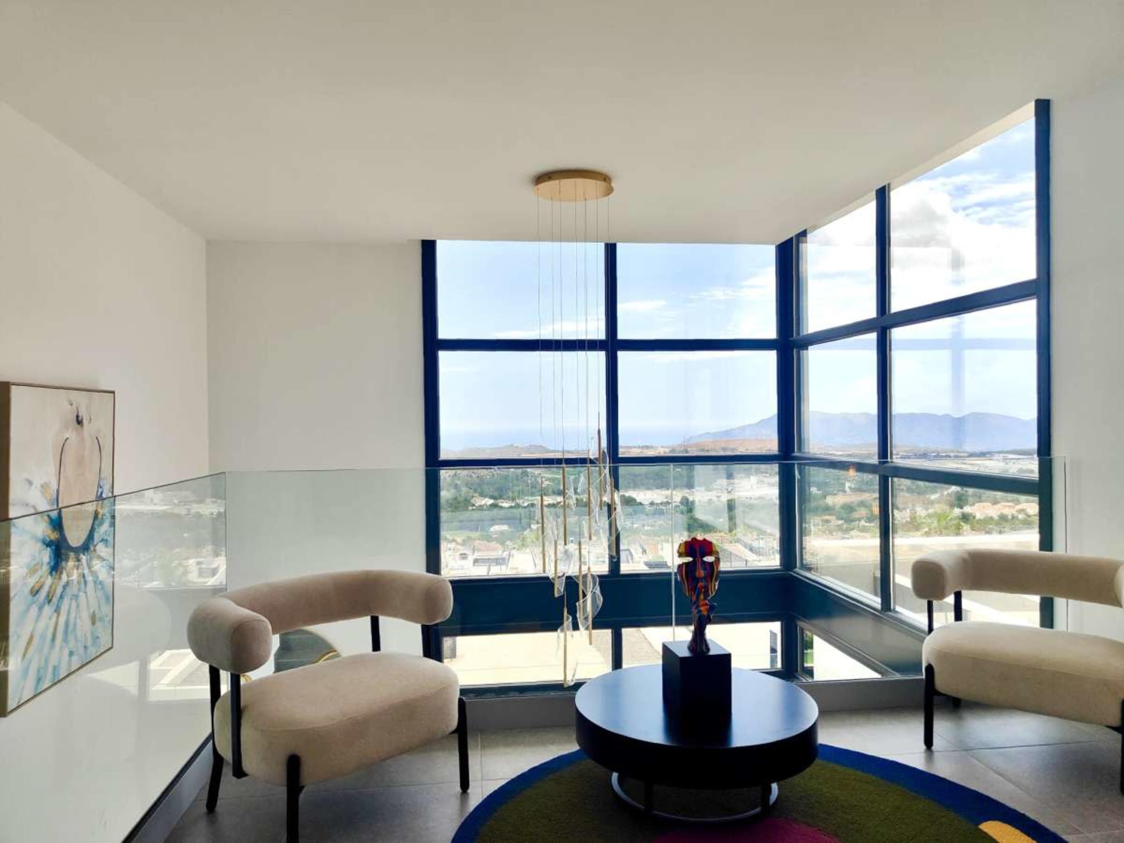 The image shows a modern interior with two cream-colored armchairs, a round black coffee table, and large windows that provide a view of the landscape outside.