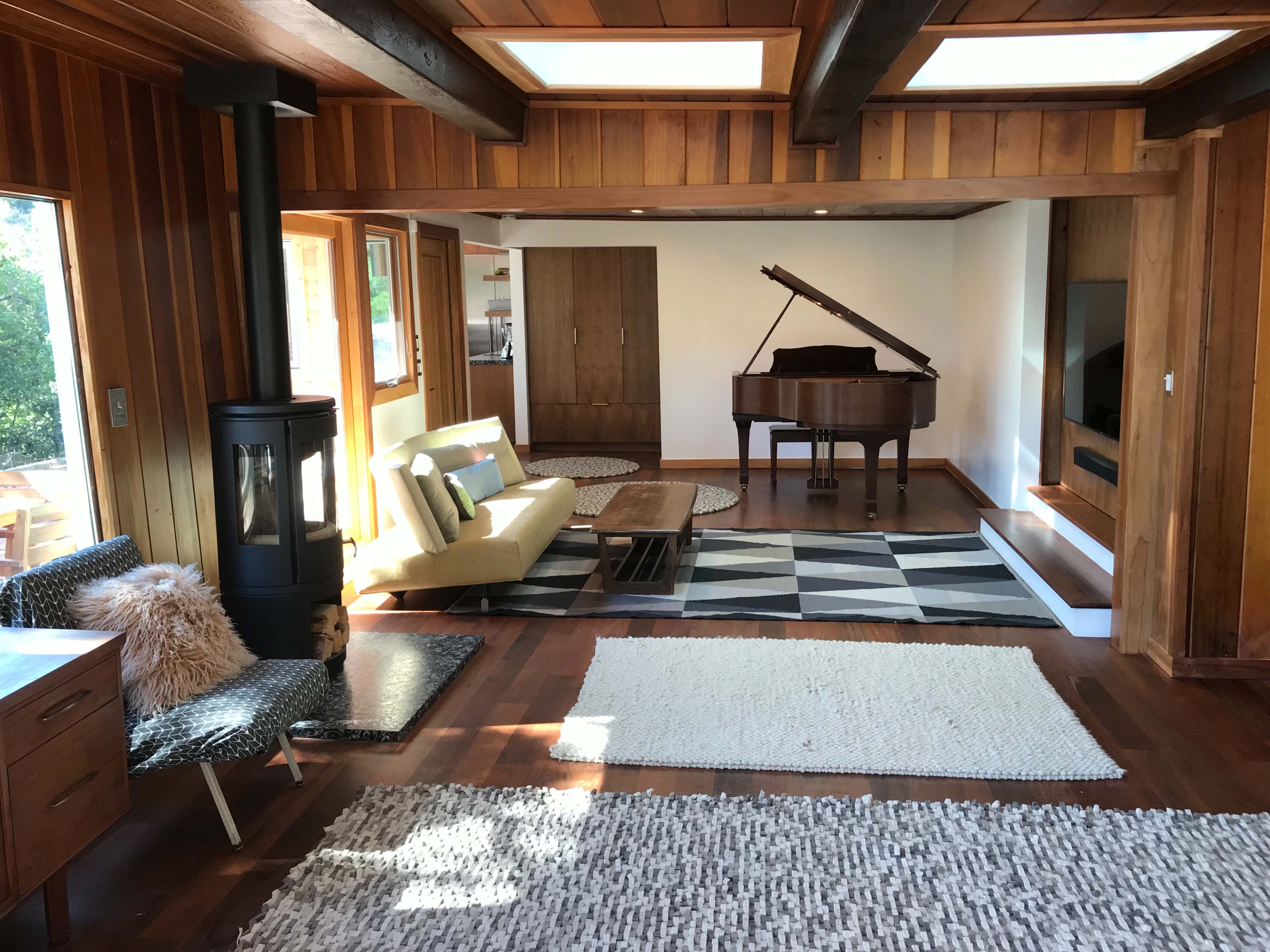 A spacious living room features a grand piano, a yellow sofa, a wooden dining table, and multiple rugs on a hardwood floor.
