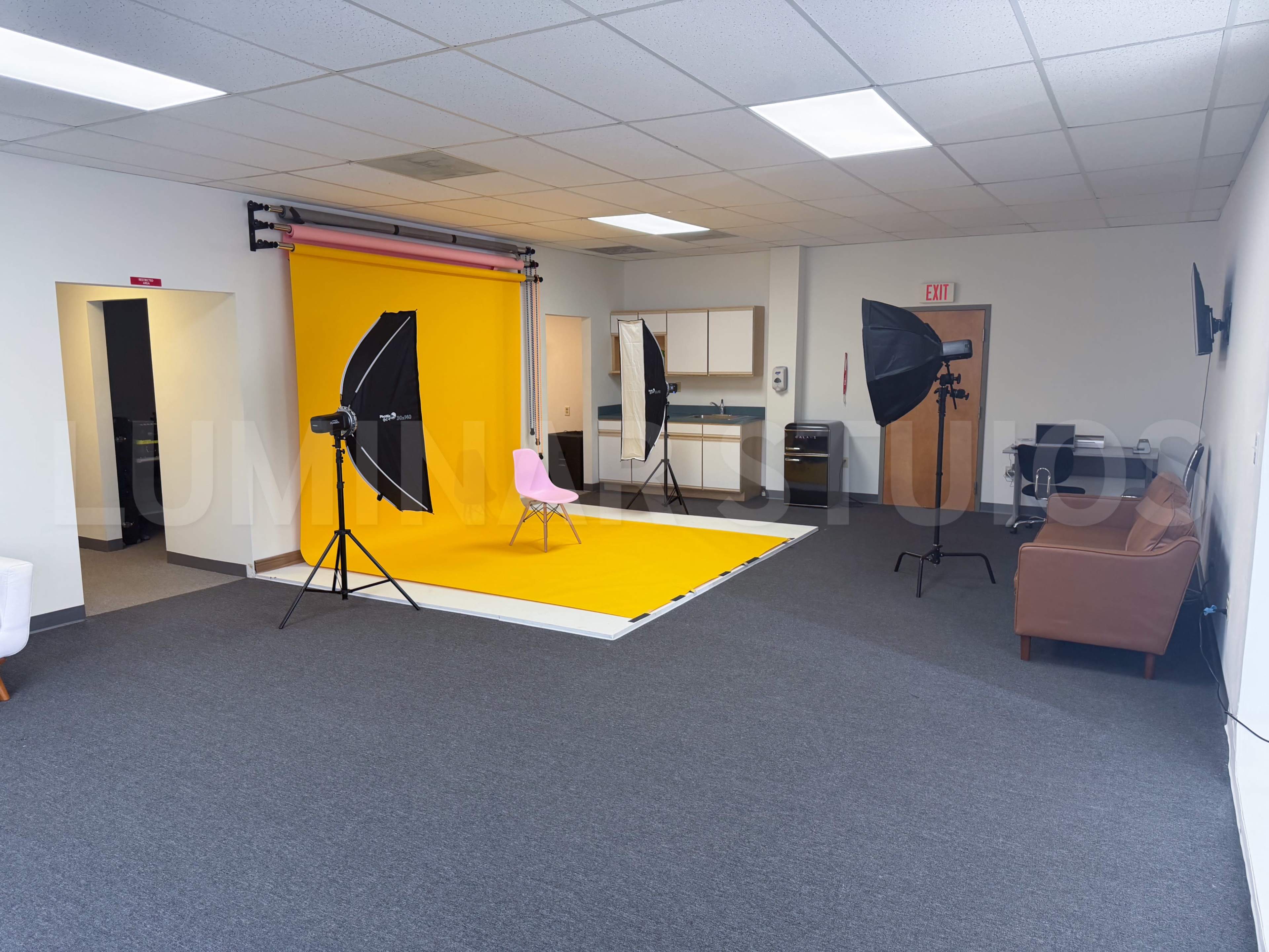 The image shows a photography studio setup, featuring a yellow backdrop, two umbrella softbox lights, a pink chair, and a brown couch in a spacious room with gray carpet.