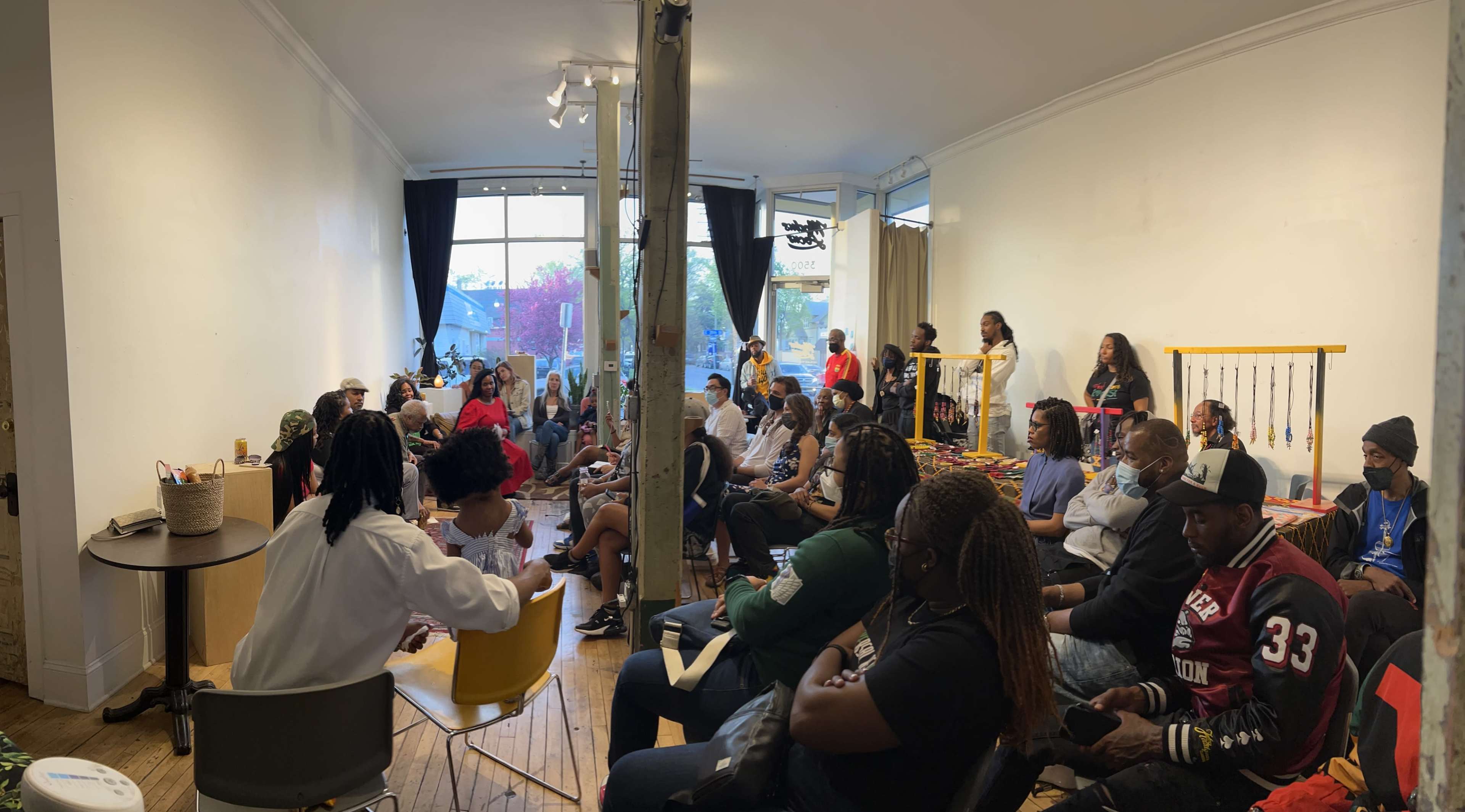 A diverse group of people is seated in a room with wooden floors, attending an event featuring displays of handmade crafts.