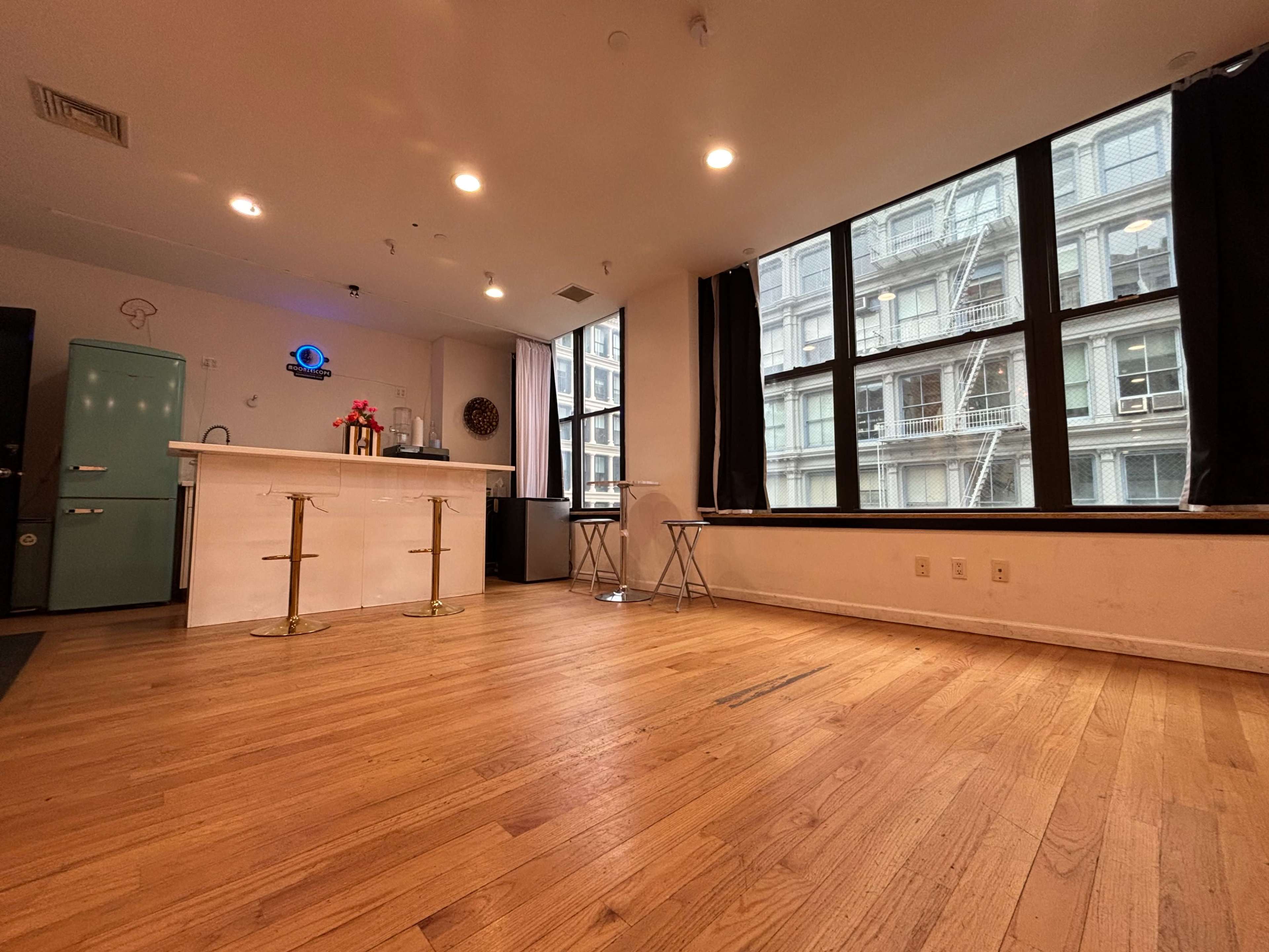 The image shows a spacious, empty room with wooden flooring, a kitchen area featuring a light green refrigerator, and large windows overlooking a cityscape.