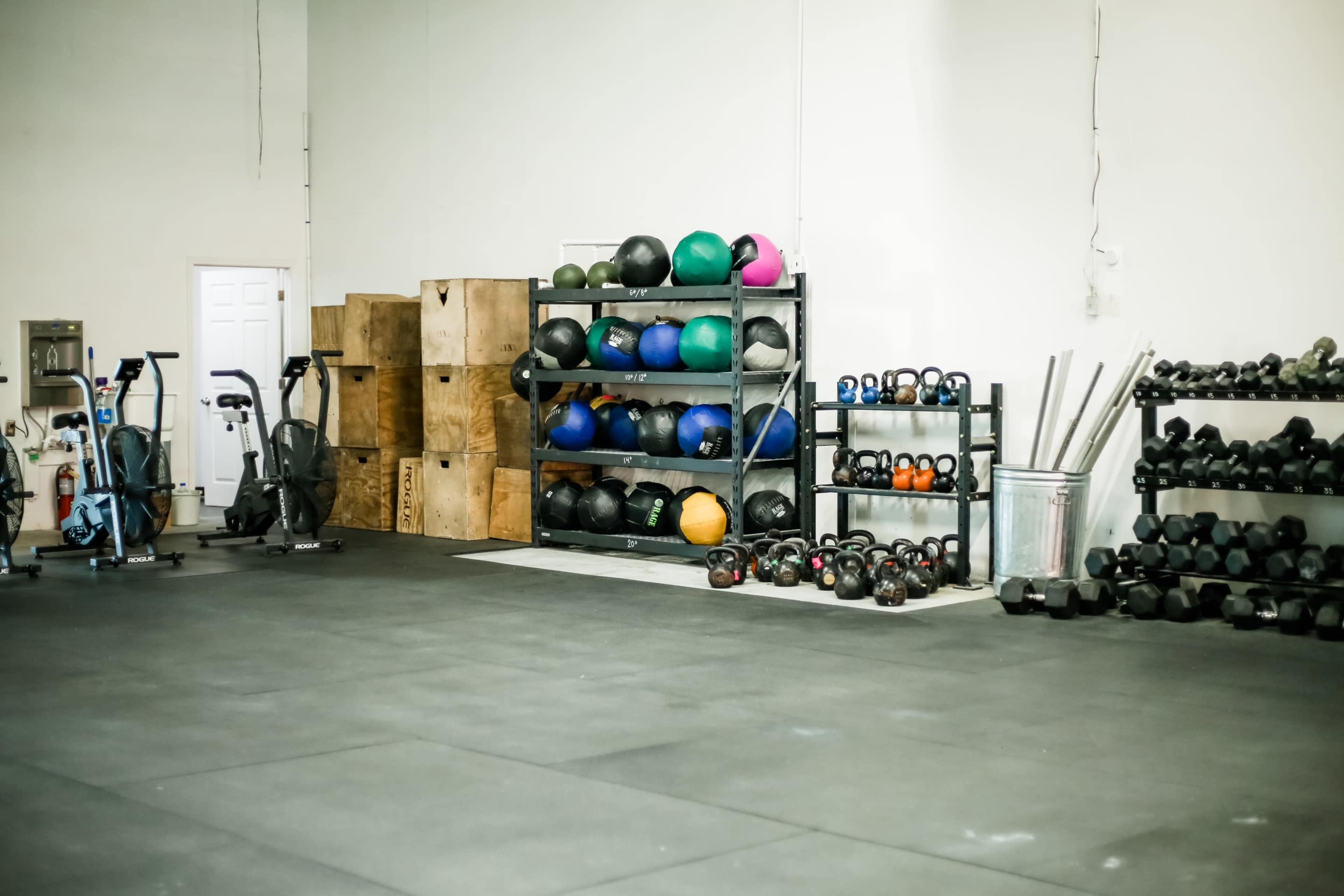 A gym interior with stacked rubber balls on shelves, various dumbbells on the floor, and workout equipment against the walls.
