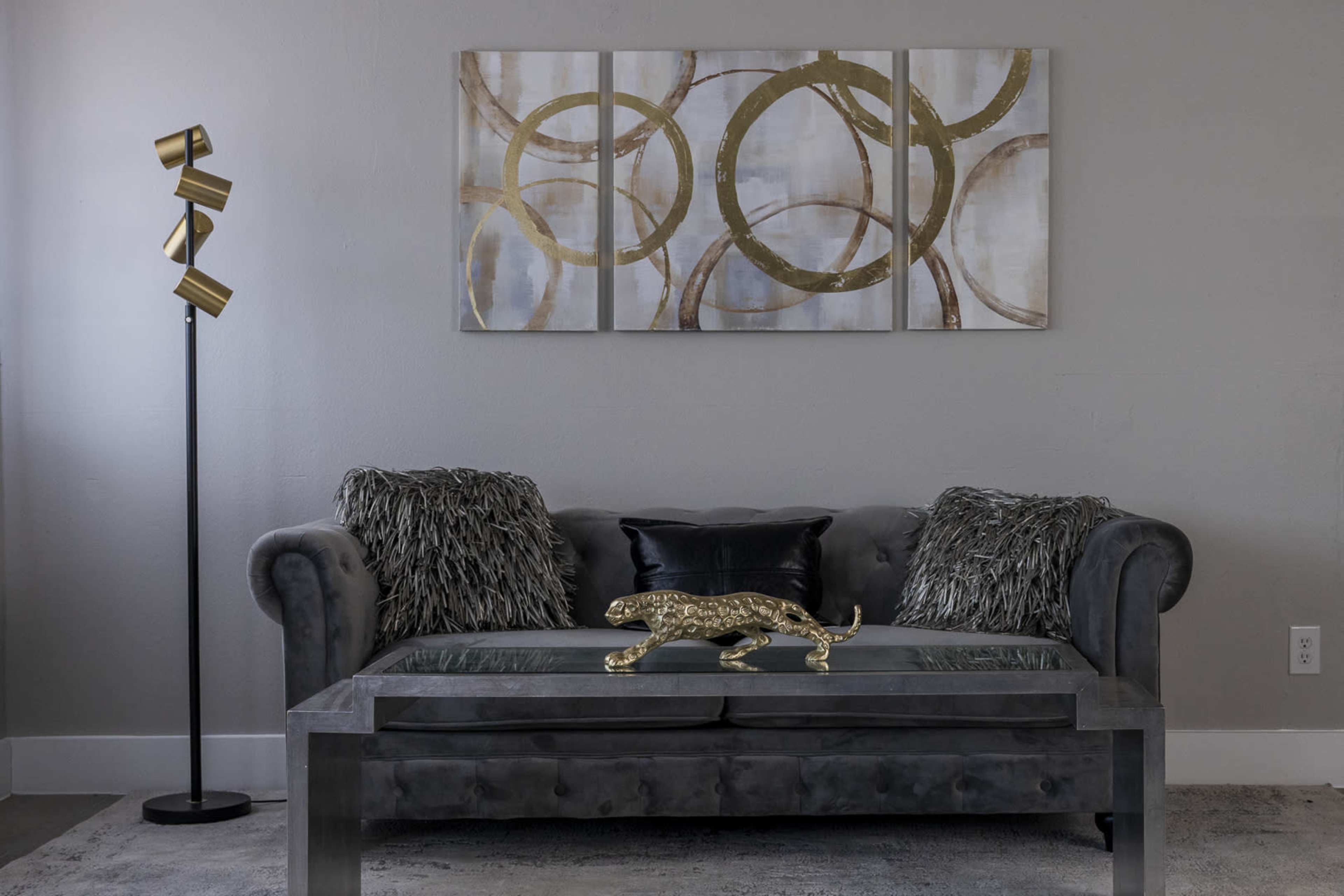 The image shows a modern living room featuring a gray sofa with decorative pillows, a contemporary coffee table with a decorative gold leopard sculpture, and a floor lamp with geometric shades.