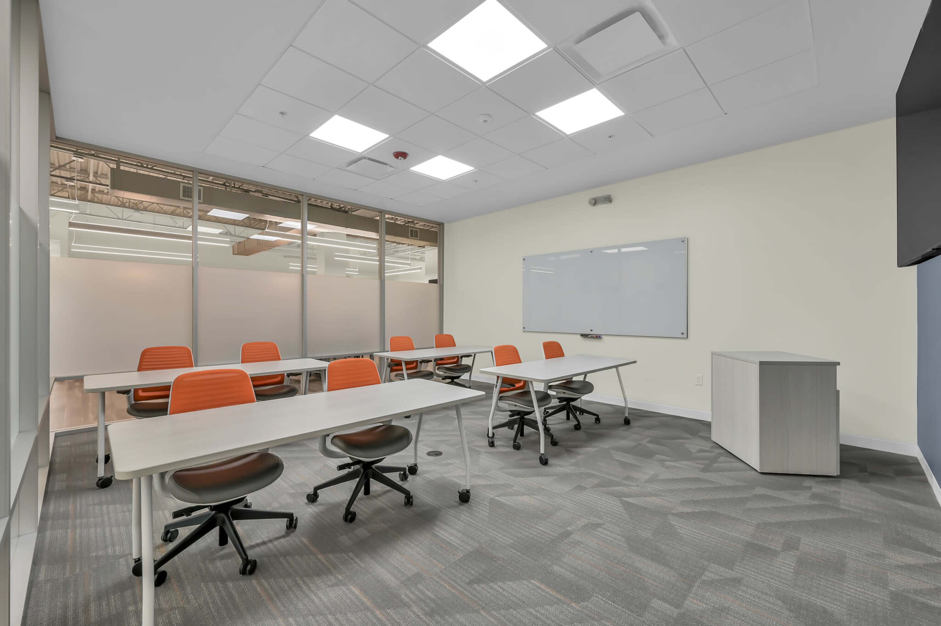 A modern classroom features several tables with orange chairs, a whiteboard, and large windows along one wall.