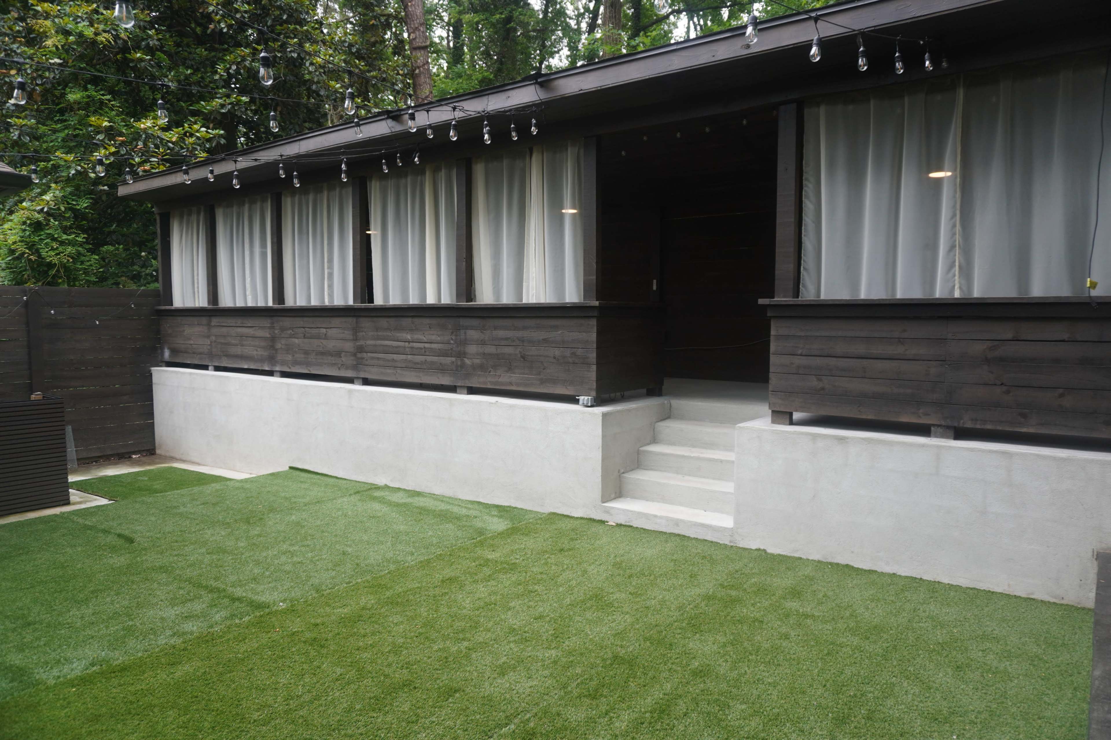 The image shows a modern wooden cabin with large windows, situated next to a green lawn and steps leading to a concrete platform.