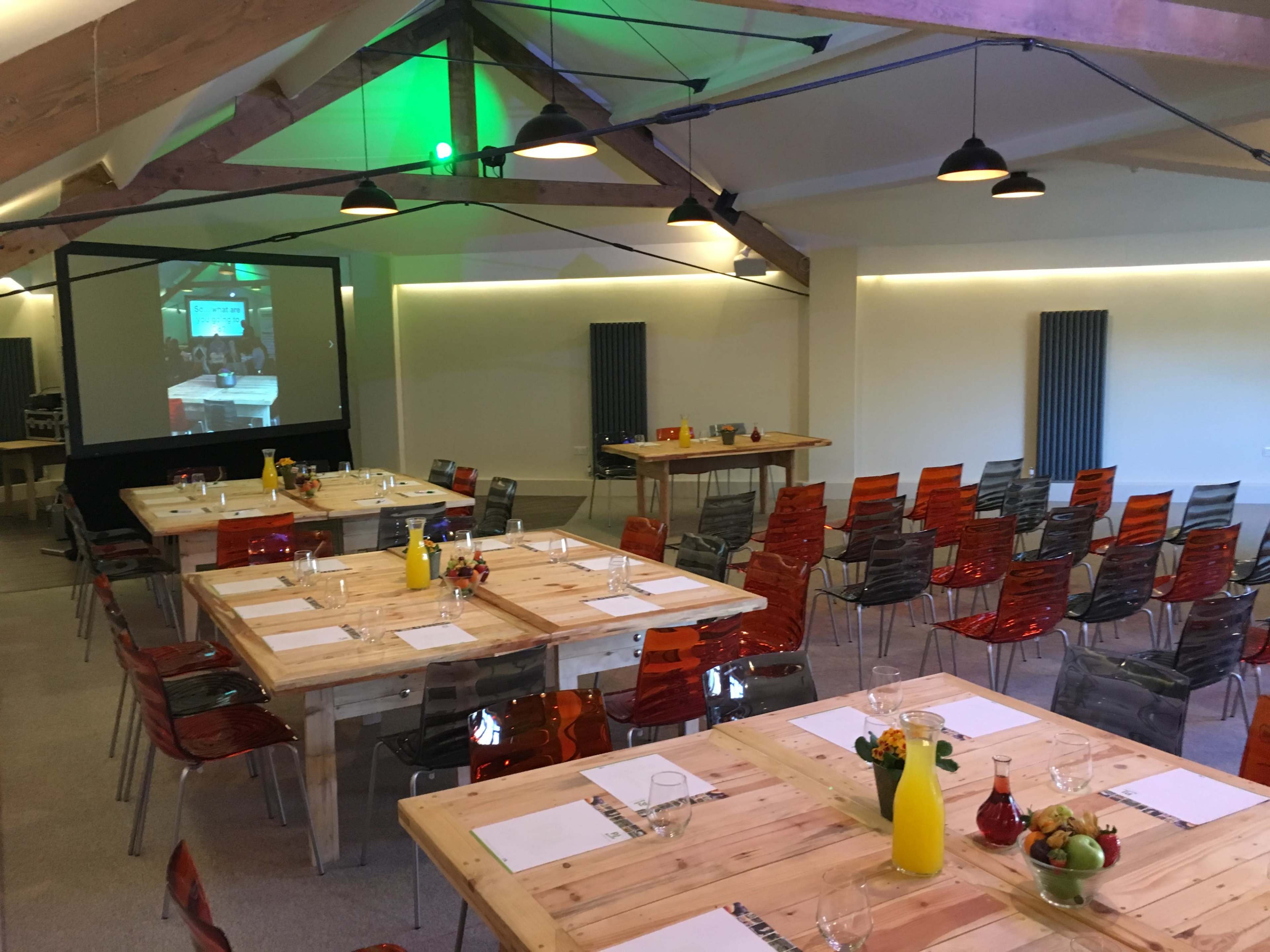 A spacious meeting room is set up with several wooden tables arranged for dining, surrounded by colorful chairs and featuring a projection screen at one end.