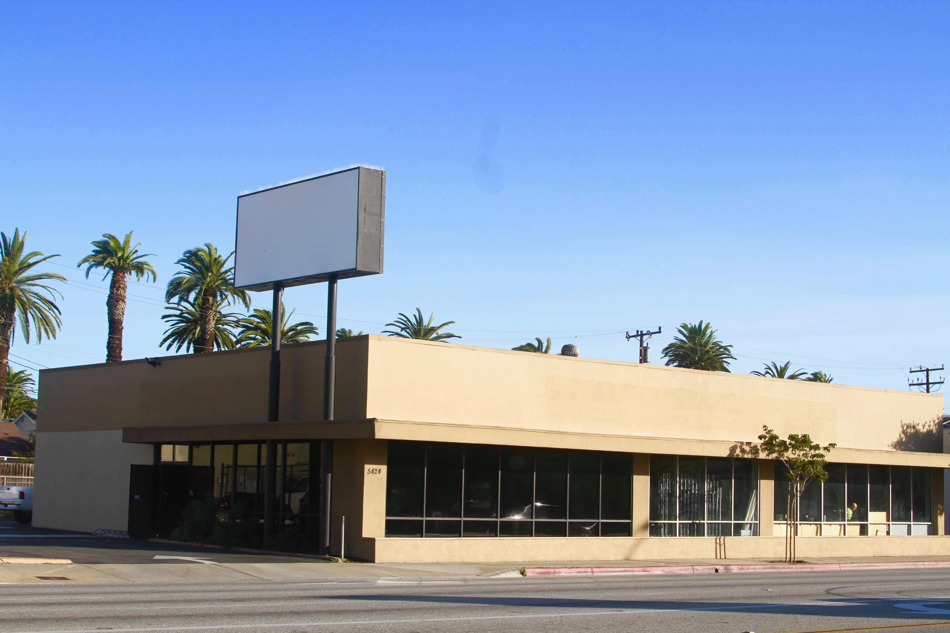 A one-story commercial building with a blank outdoor sign is surrounded by palm trees on a sunny day.