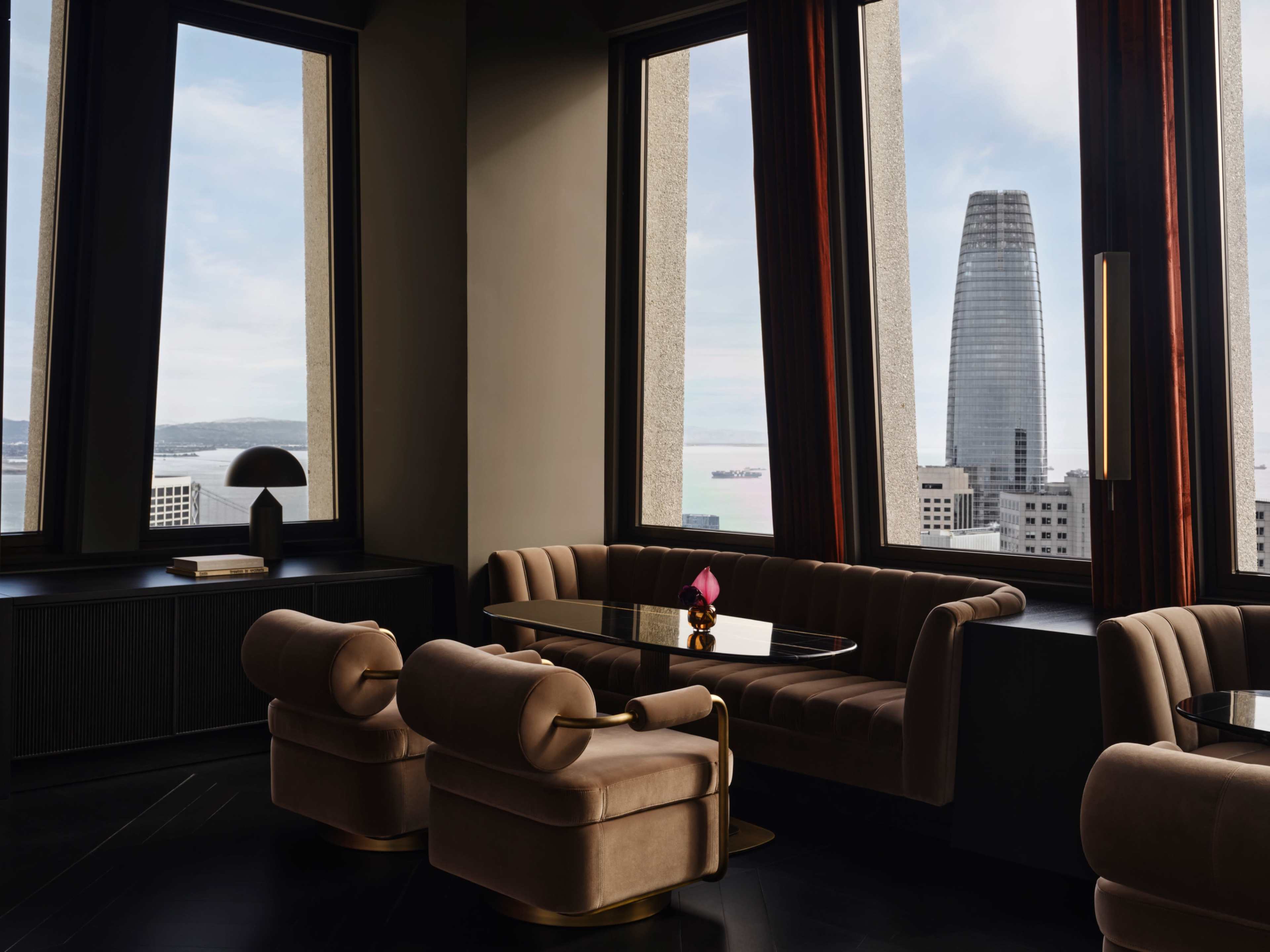 The image shows a modern lounge area with a plush sofa and chairs, featuring large windows that overlook a cityscape with a tall, distinctive building in the background.