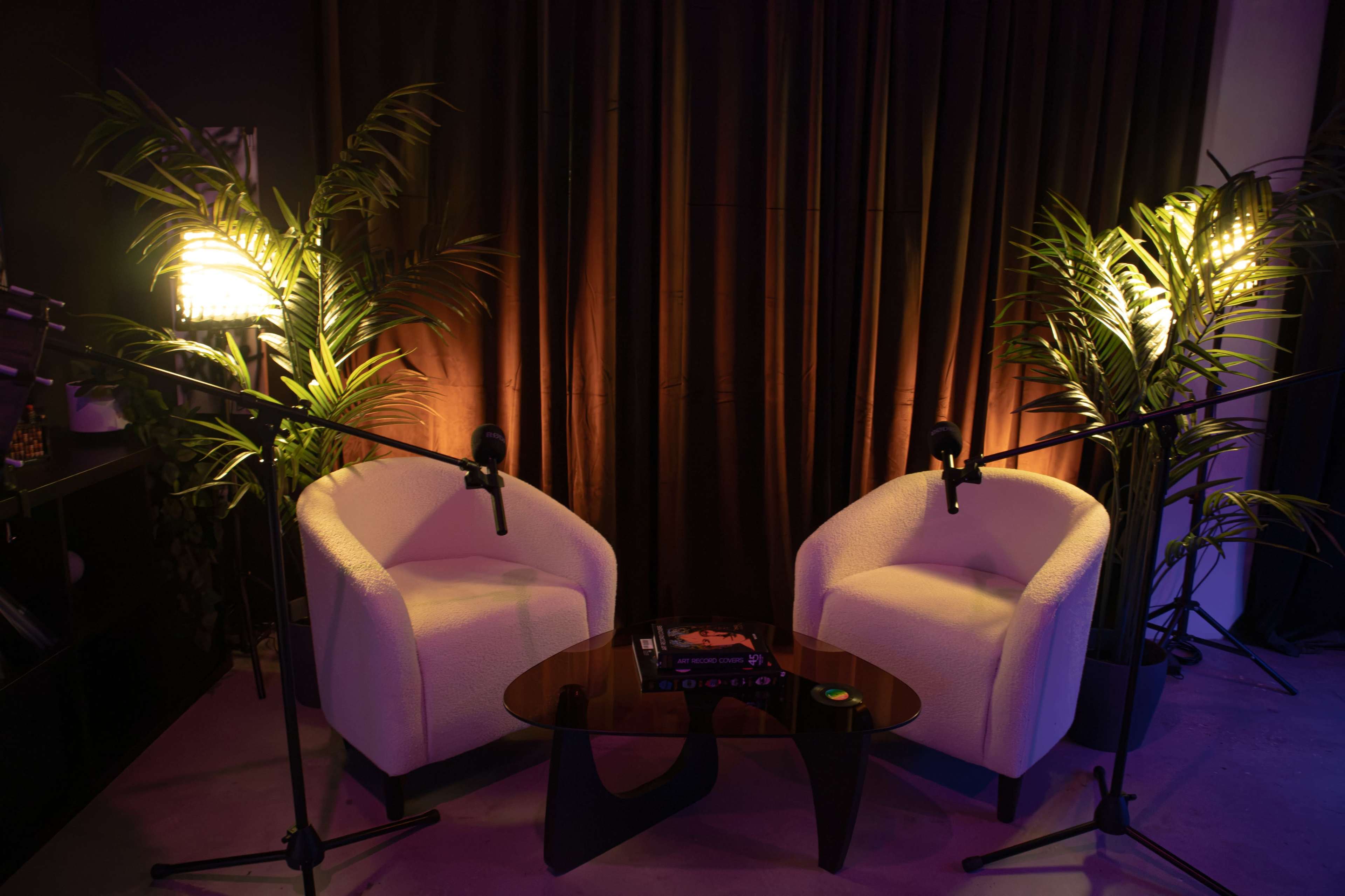 The image shows a cozy seating area featuring two white armchairs, a round wooden coffee table, and two floor lamps, all surrounded by potted plants and a dark curtain backdrop.