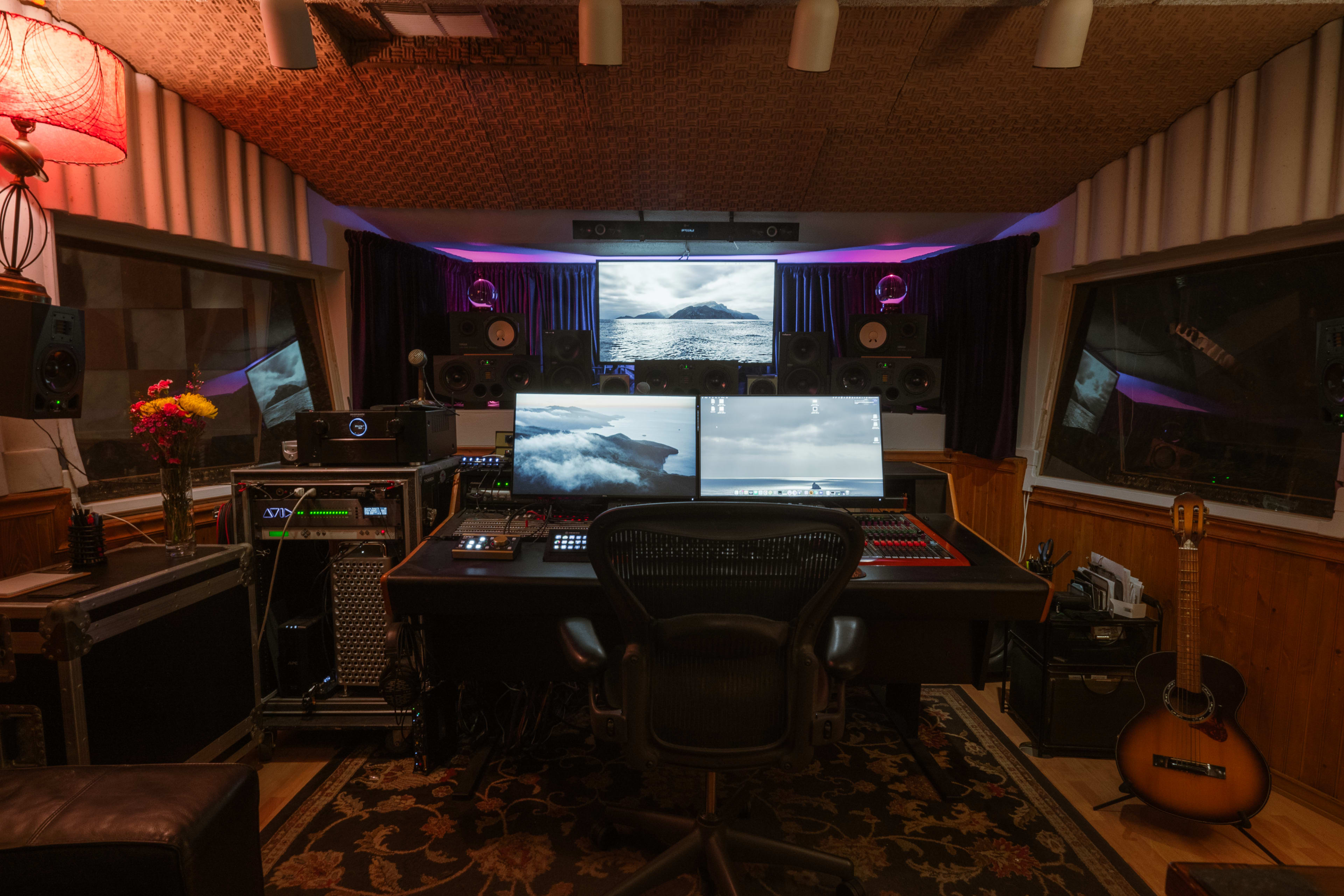 The image shows a music studio with a central workstation featuring two large screens, surrounded by audio equipment, speakers, and a guitar resting on a chair.