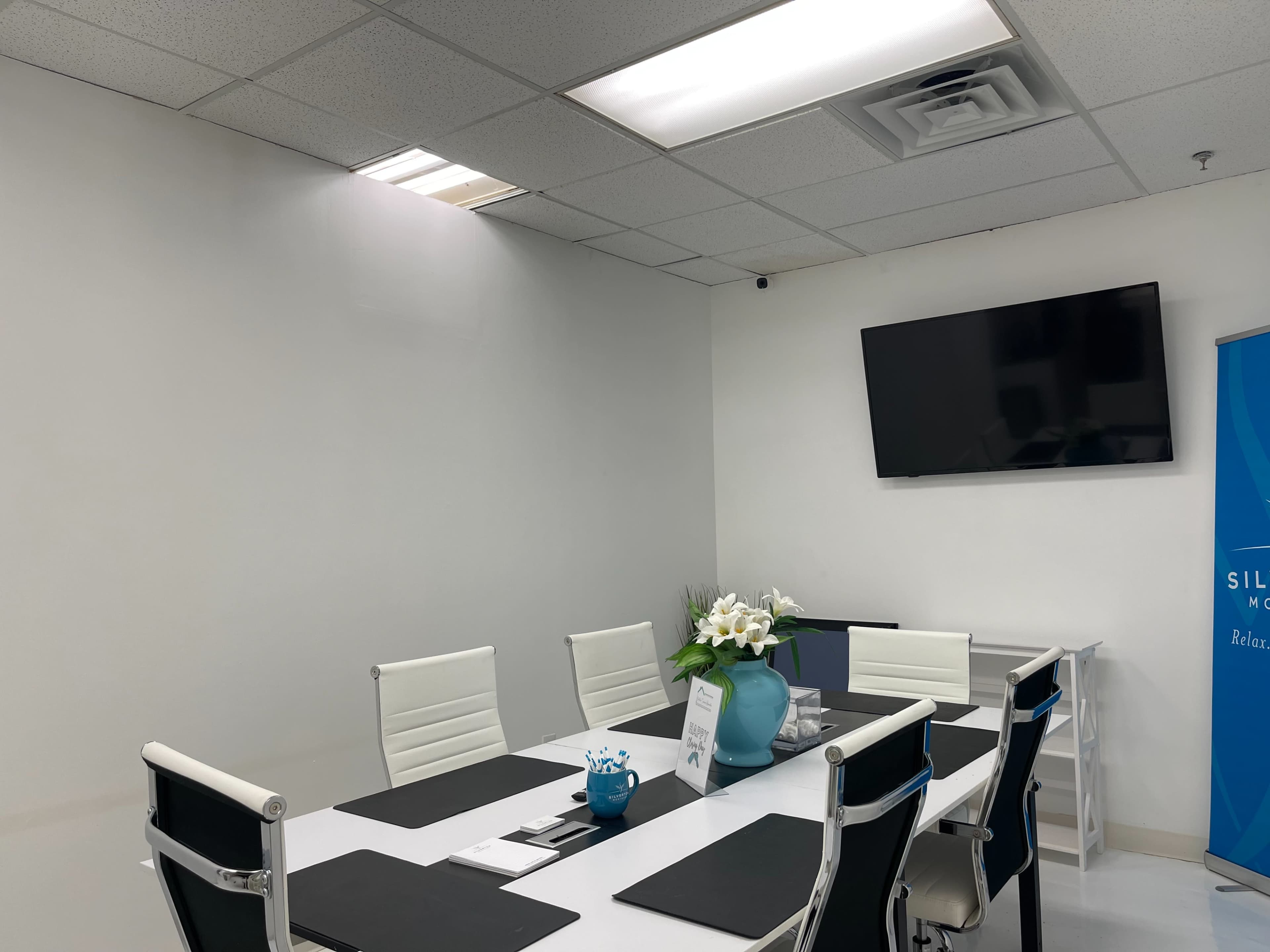 A modern conference room features a rectangular table surrounded by white chairs, with a television mounted on the wall and a vase of flowers in the center.