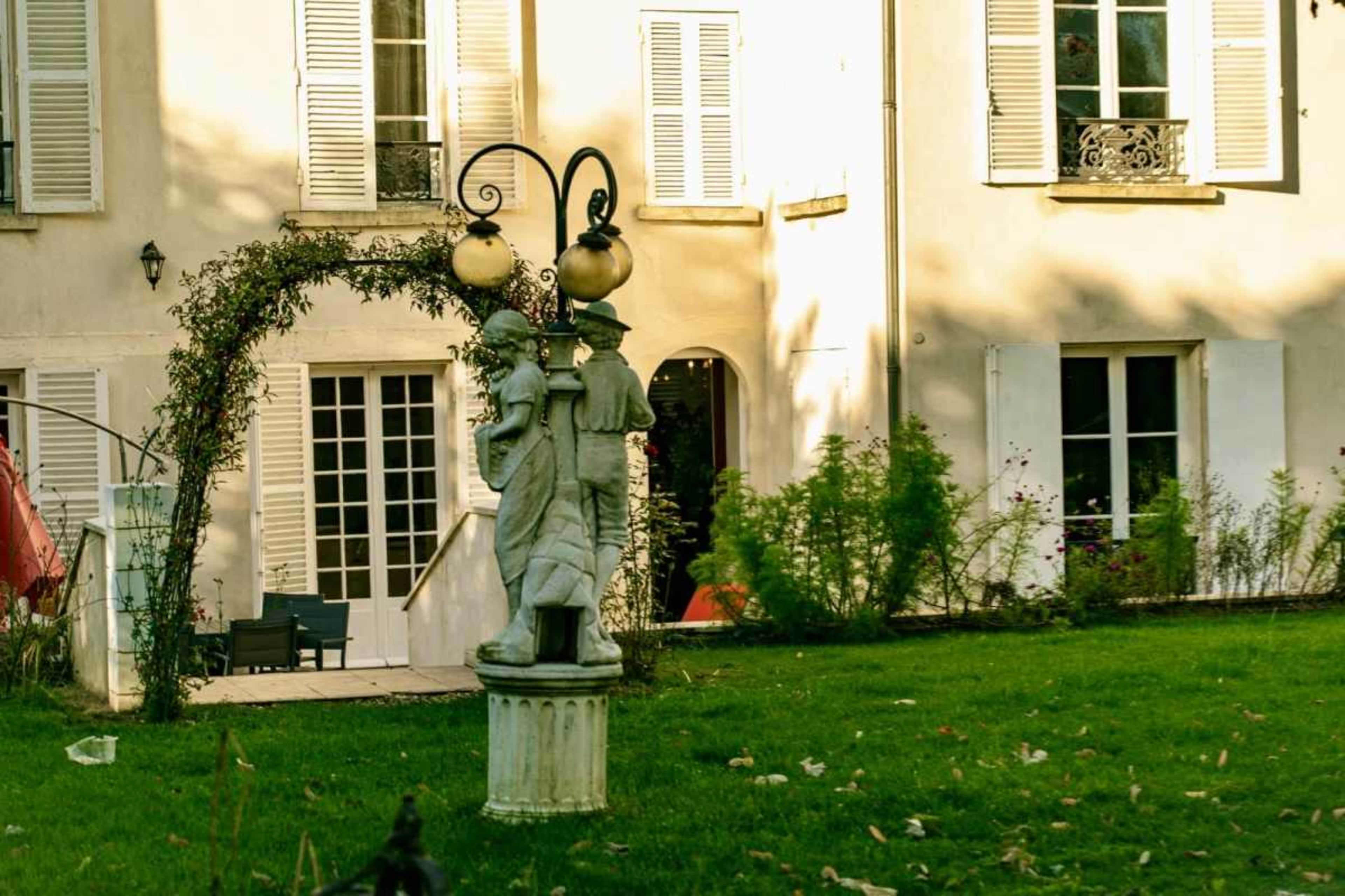A statue holding a lantern is situated in a garden in front of a house with white shutters and a large arched entrance adorned with greenery.