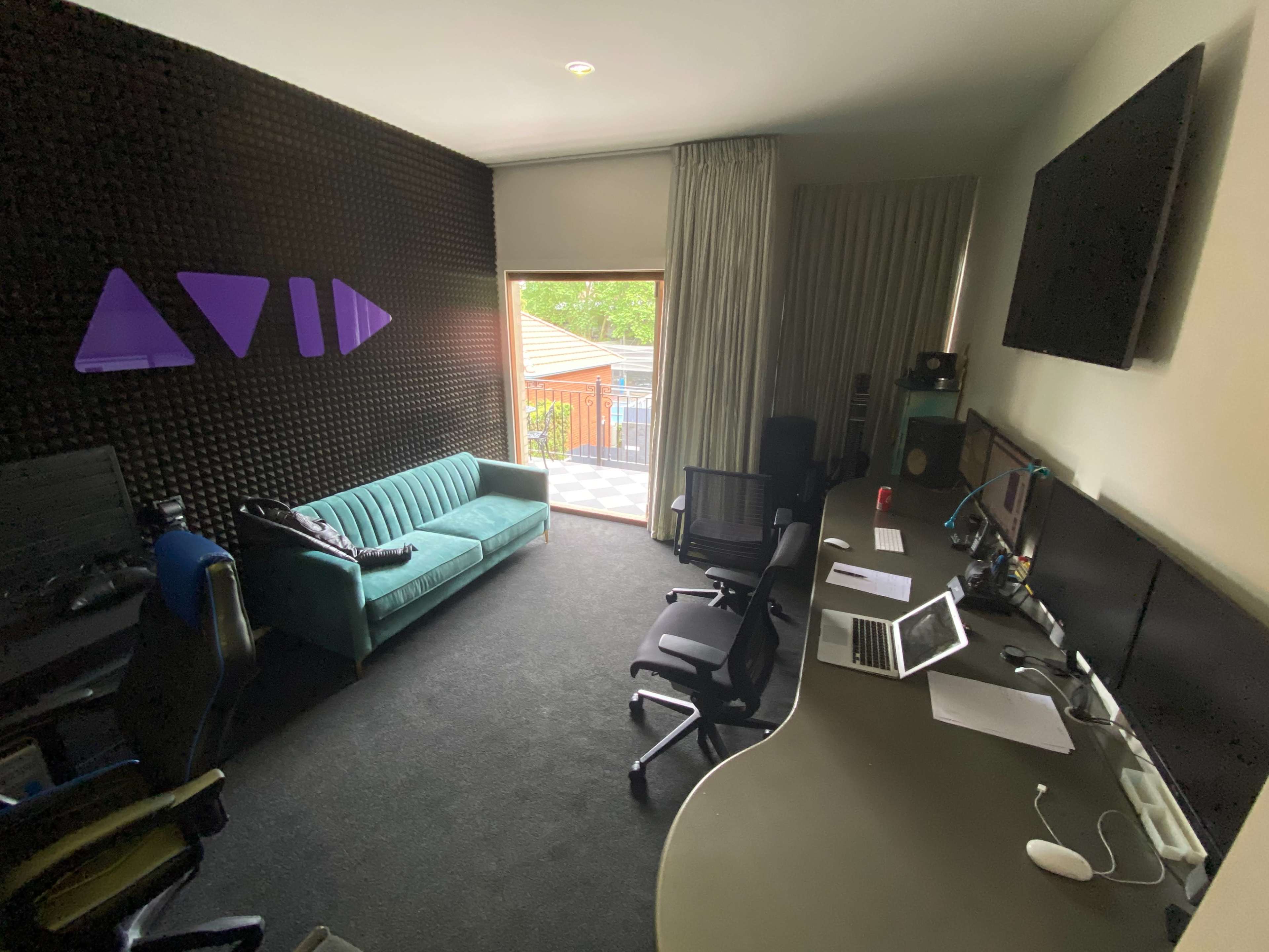 The image shows a modern office space with a turquoise couch, dual workstations with computers, and a large window leading to an outdoor deck.