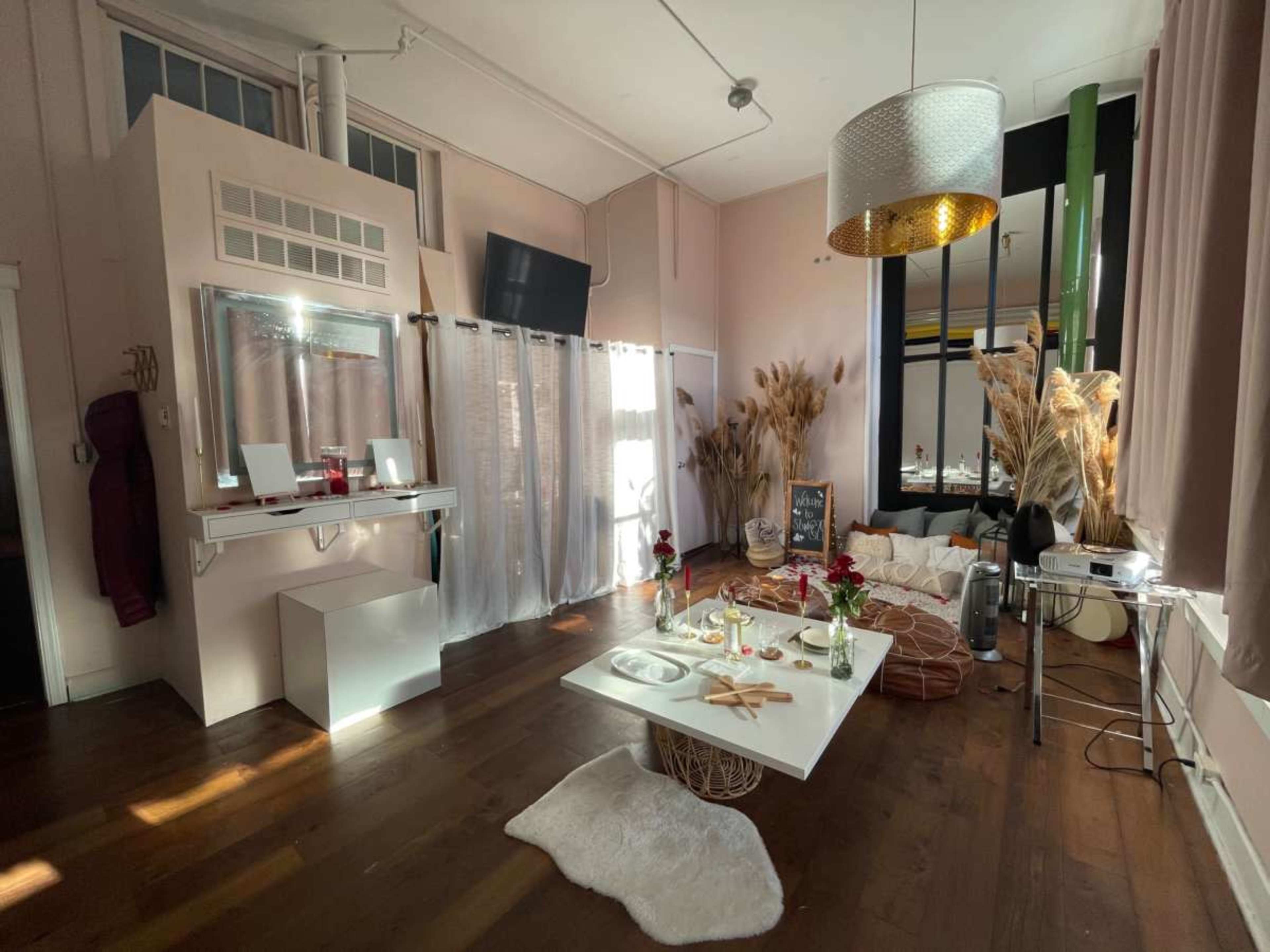 The image shows a warmly lit indoor space with a mix of modern and cozy decor, featuring a low table surrounded by cushions, plants, and various decorative elements.