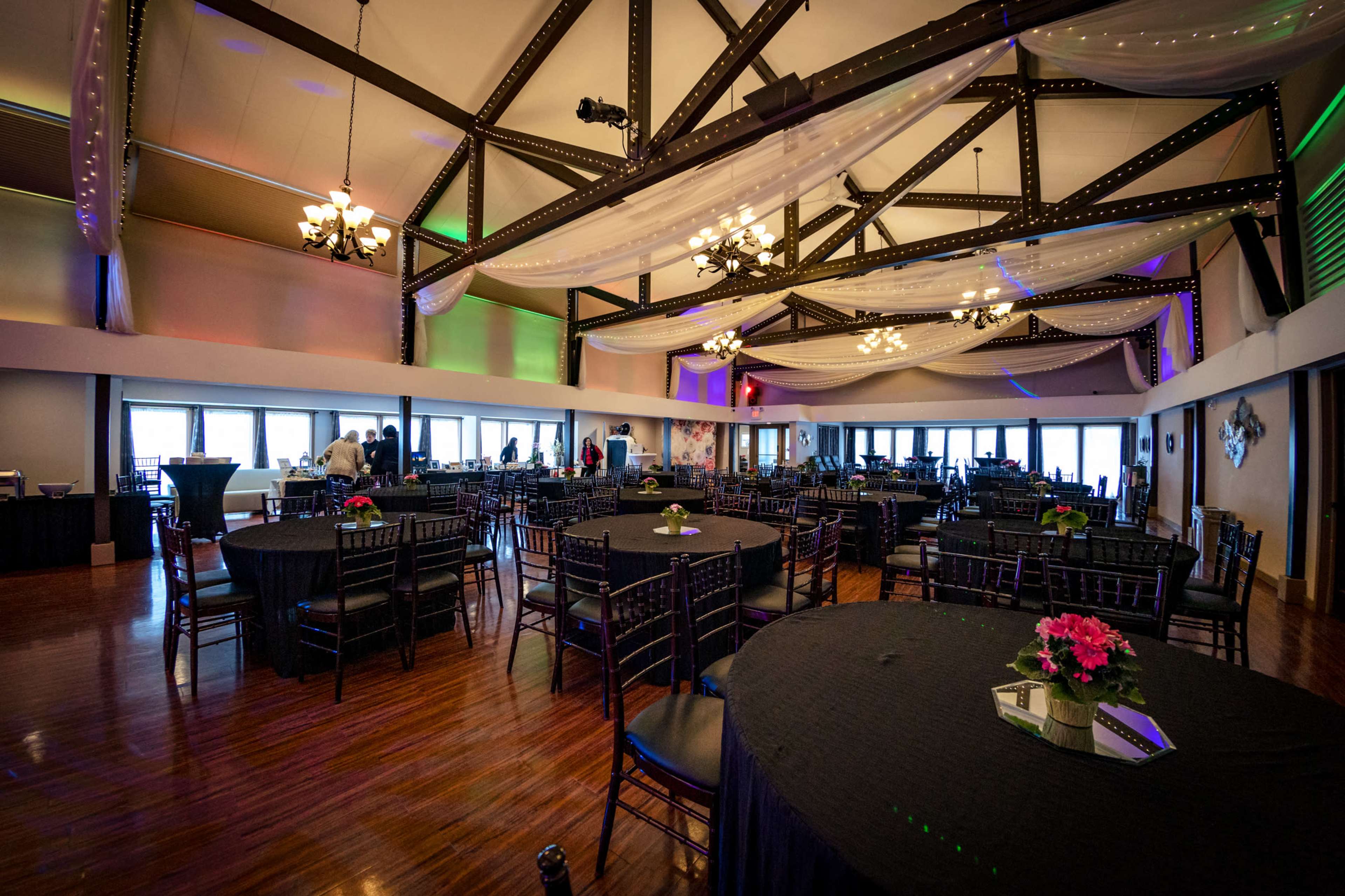 An elegantly arranged banquet hall with black tables, chairs, and floral centerpieces, featuring high ceilings and decorative lighting.