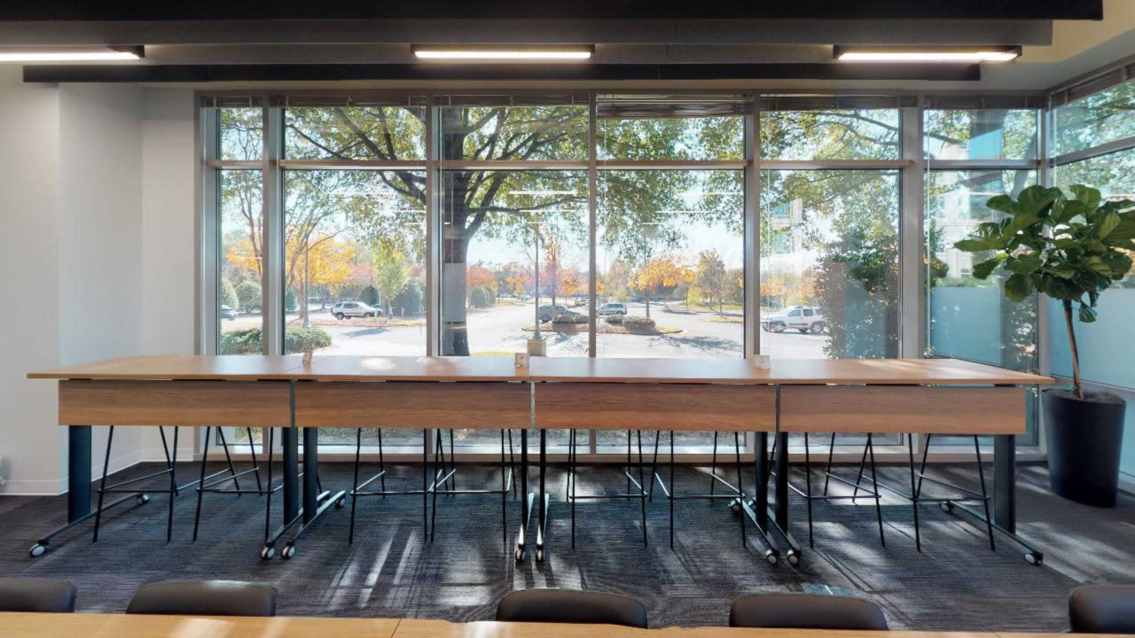 A row of empty tables is positioned against large windows overlooking an outdoor scene with trees and parked cars.